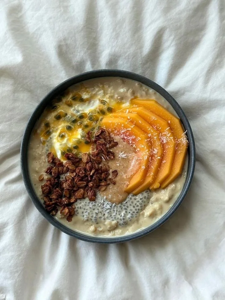A bowl of oatmeal topped with passion fruit pulp, sliced mango, toasted coconut flakes, and chopped nuts.