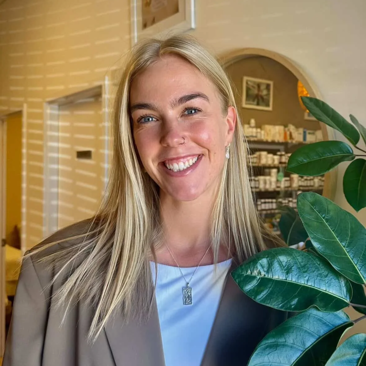 A smiling blonde woman with blue eyes wearing a beige blazer, a white top, a silver necklace, and earrings, standing indoors near a green plant with large leaves, with shelves of bottles and a picture in the background.