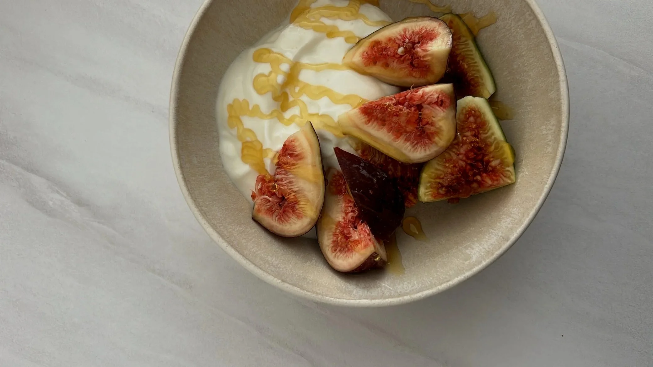 A bowl containing sliced figs, Greek yogurt topped with honey and a drizzle of honey over the yogurt, on a light gray surface.