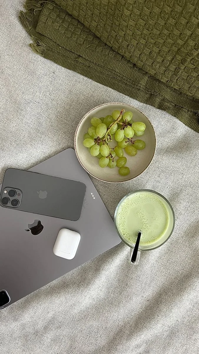 Close-up of a table with a gray iPad, a white AirPods case, a silver iPhone, a bowl of green grapes, and a glass of green juice with a straw. A green textured fabric is partially visible at the top of the image.