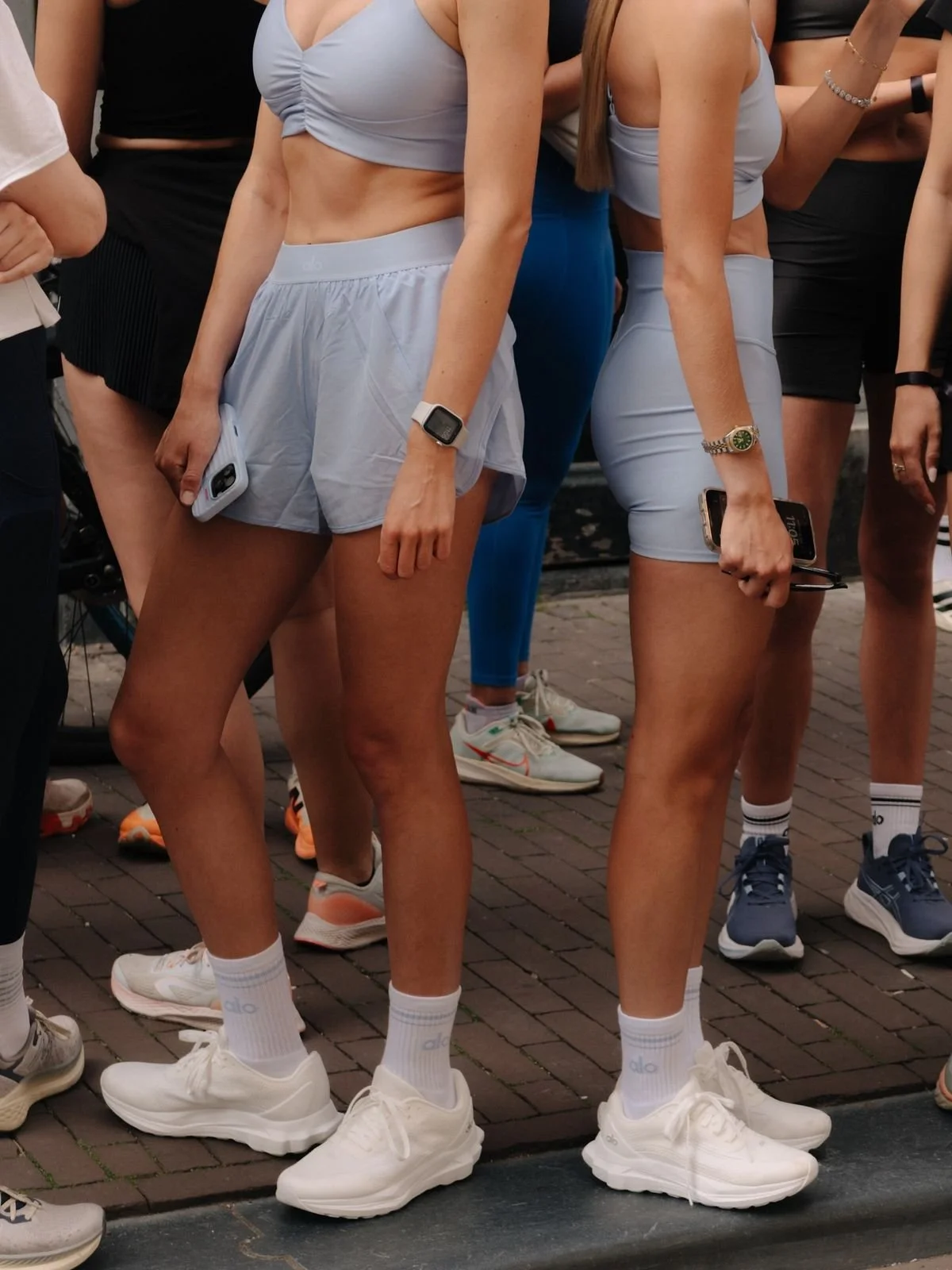 Group of women in athletic clothing, holding phones, standing on brick pavement, some wearing white sneakers and white socks, in an outdoor setting.