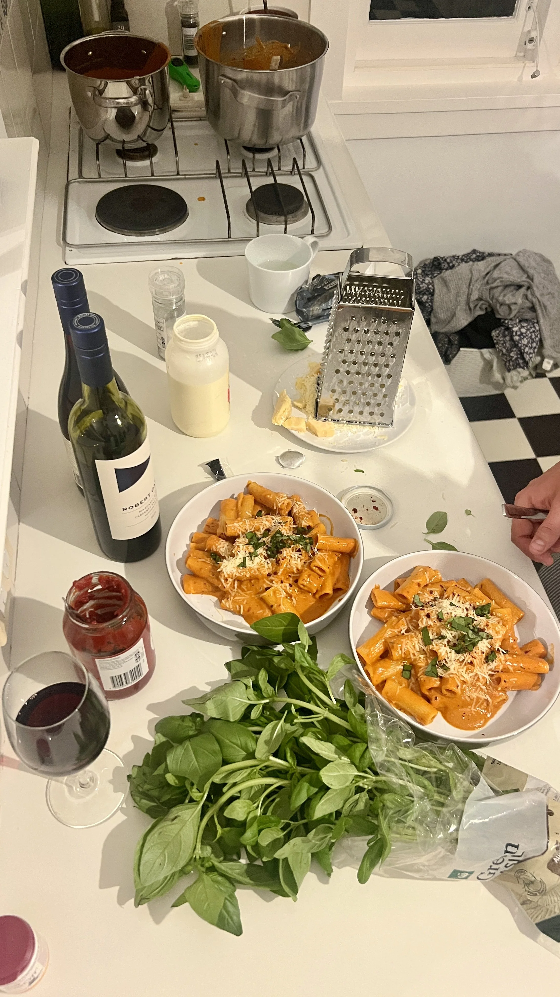 A kitchen countertop with two bowls of pasta in tomato sauce, two glasses of red wine, bottles of wine, a jar of tomato sauce, a basil plant, a box of grated cheese, and various kitchen items. There are two pots on the stove, one with tomato sauce and the other with cooked pasta.