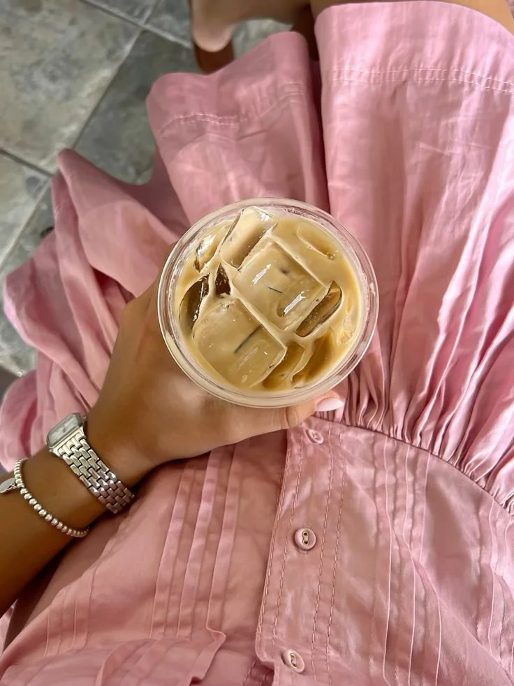 A person wearing a pink dress holding a cup of iced coffee with ice cubes, viewed from above.