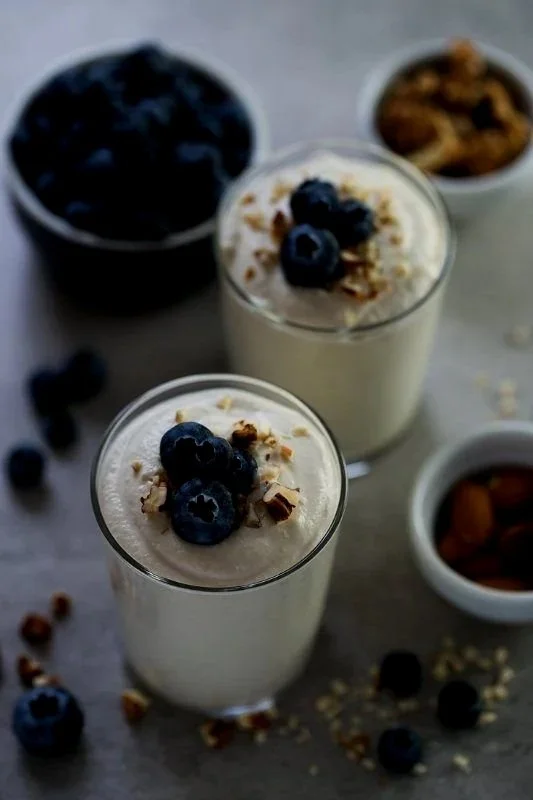 Two glasses of yogurt topped with blueberries and granola, with small bowls of blueberries, granola, and almonds in the background.