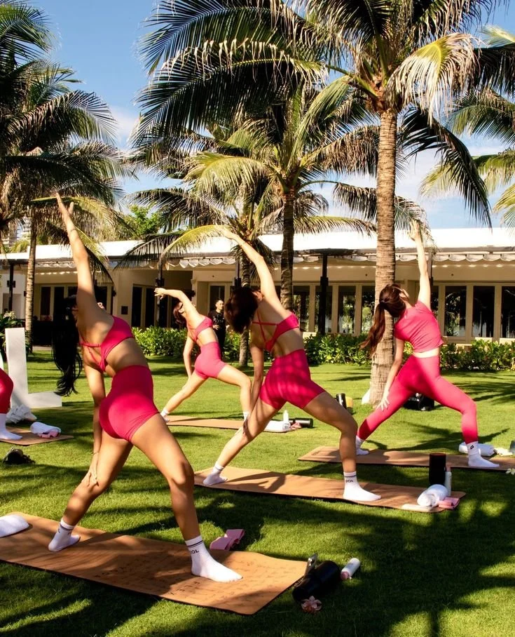 Group of women practicing yoga outdoors on yoga mats beneath palm trees on a sunny day.
