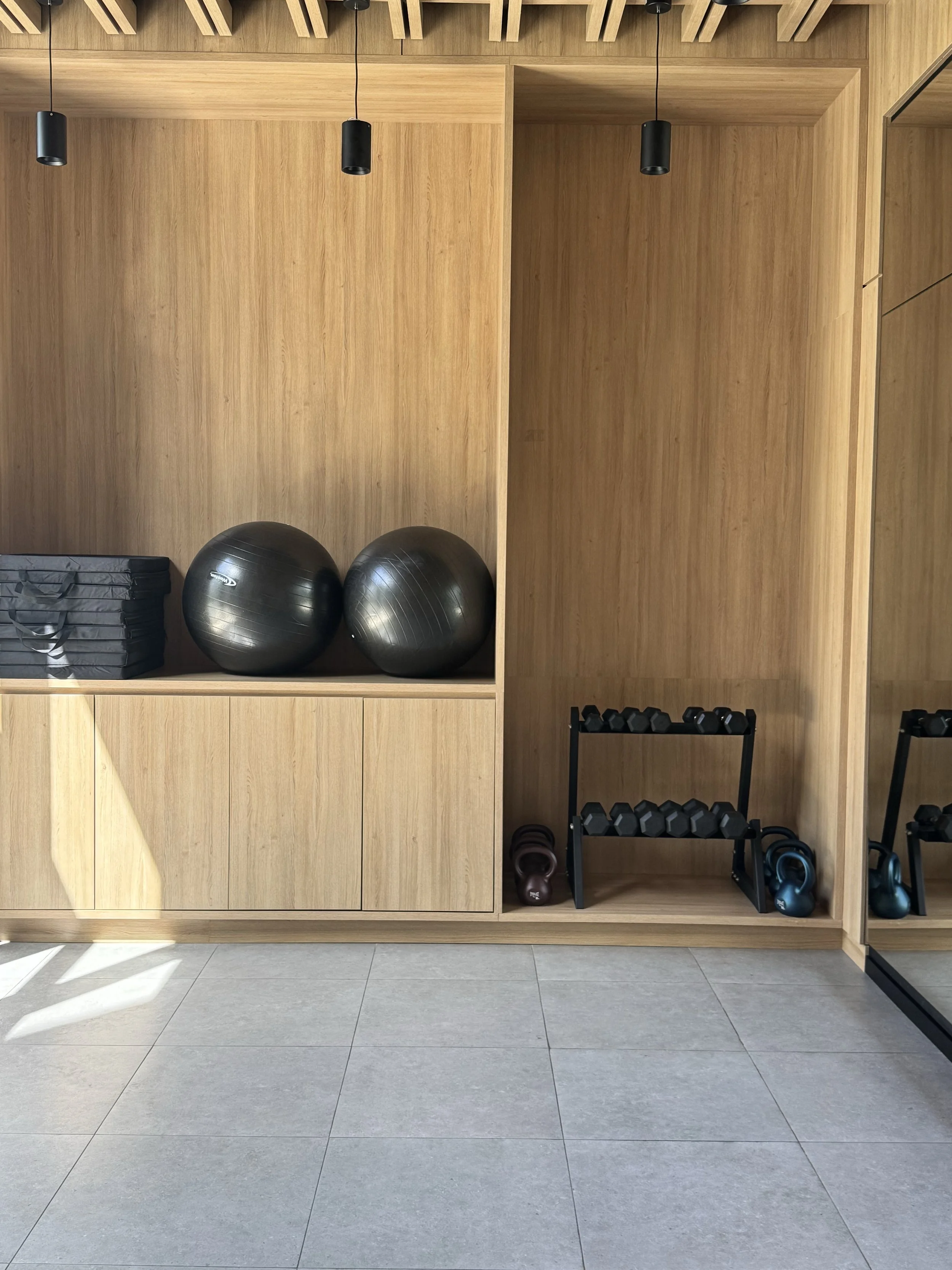 Workout equipment including two black exercise balls, a set of black dumbbells on a rack, kettlebells, and black mats stored on wooden shelves in a fitness room.