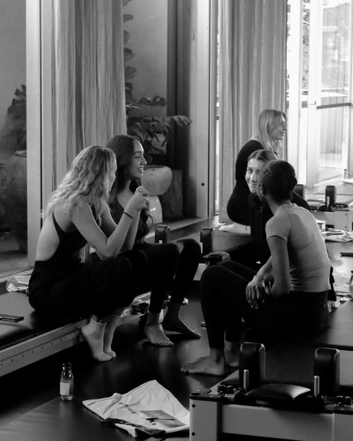 Five women sitting and talking inside a room with large windows and curtains, some on a bench and some on a table, with various objects including bottles, phones, and papers around them.