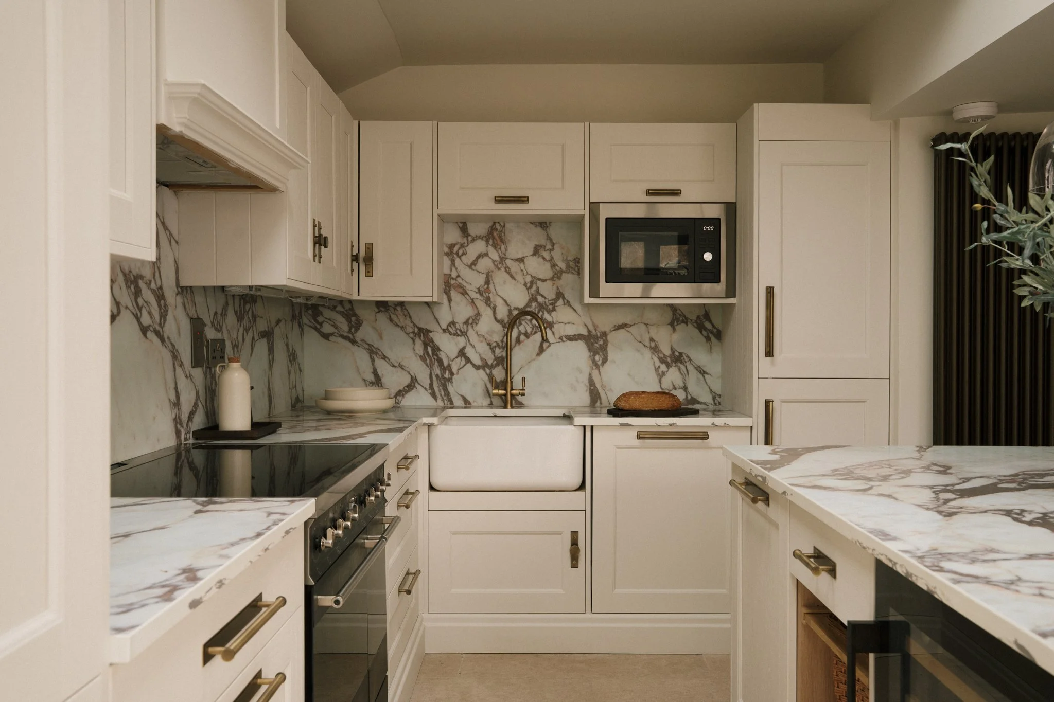 Modern kitchen with white cabinetry, marble countertops and backsplash, gold hardware, a black stove, microwave, and a farmhouse sink.