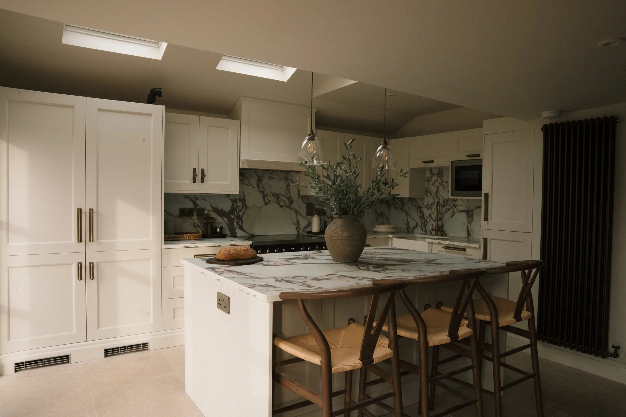 Modern kitchen with white cabinetry, marble backsplash, and a matching marble island. The island has a large vase with greenery, two pendant lights, and three wooden barstools. Skylights and a black radiator are also visible.