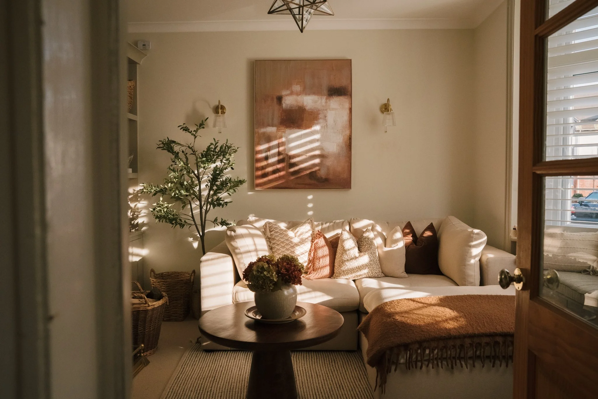  cozy living room with a white L-shaped sofa, multiple patterned pillows, a round wooden coffee table, a potted plant, woven baskets, and sunlight streaming through window blinds creating striped shadows