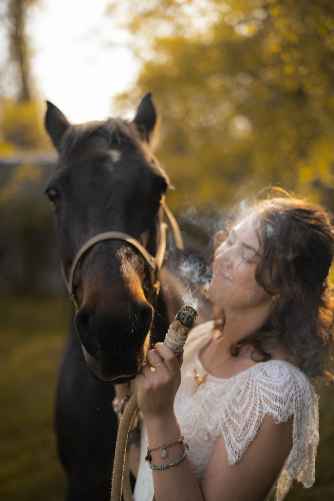 Woman in white lace dress holding smudge stick near horse, outdoors, with sunlight filtering through trees.