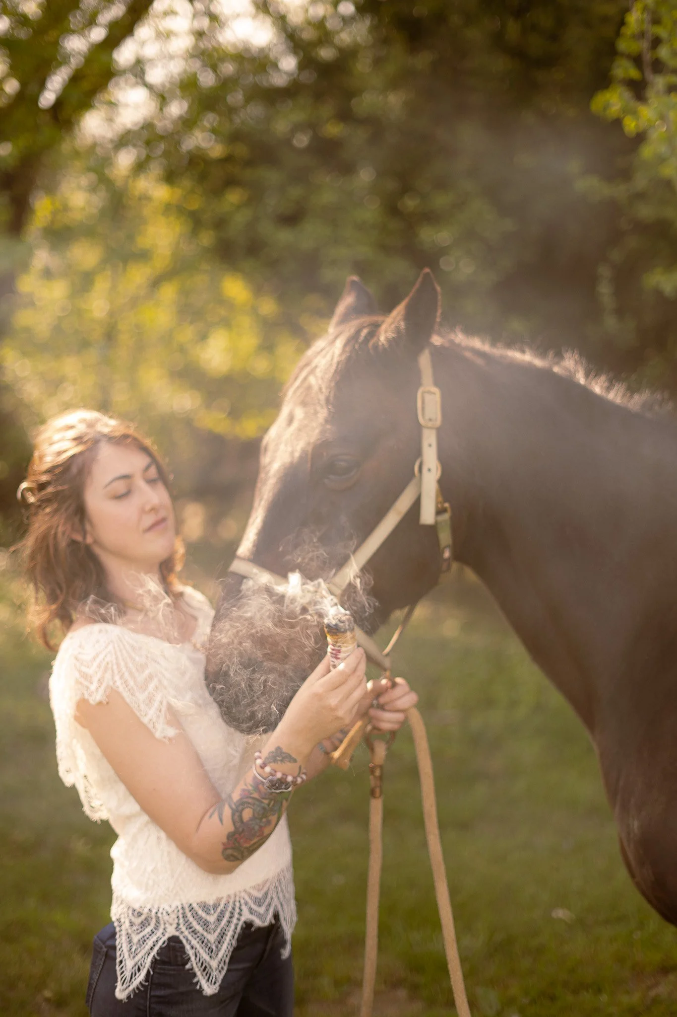 Woman with tattoo holding smoke bundle near horse in grassy area
