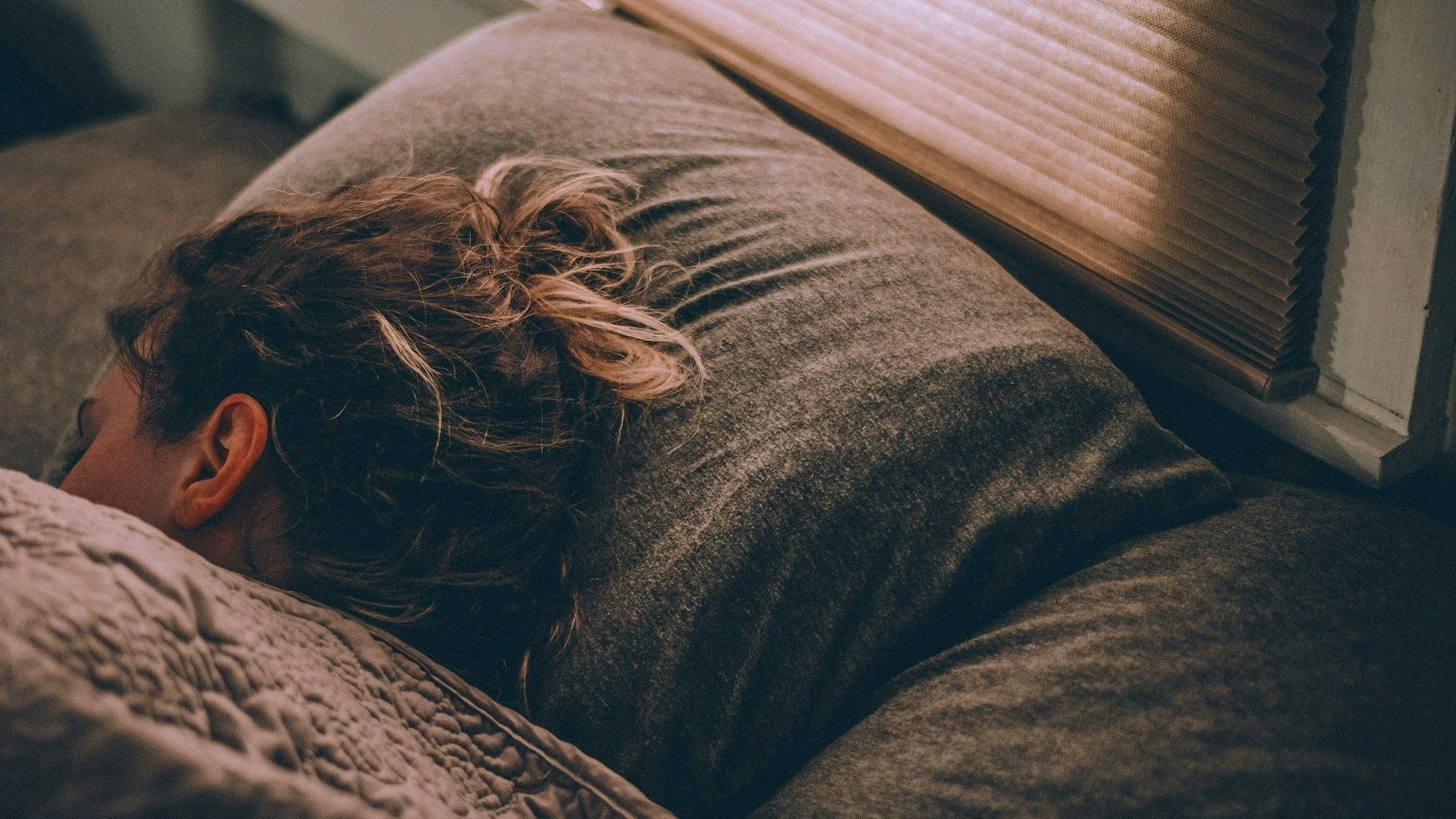Person sleeping with head on a pillow near a window with blinds, covered with a gray blanket.