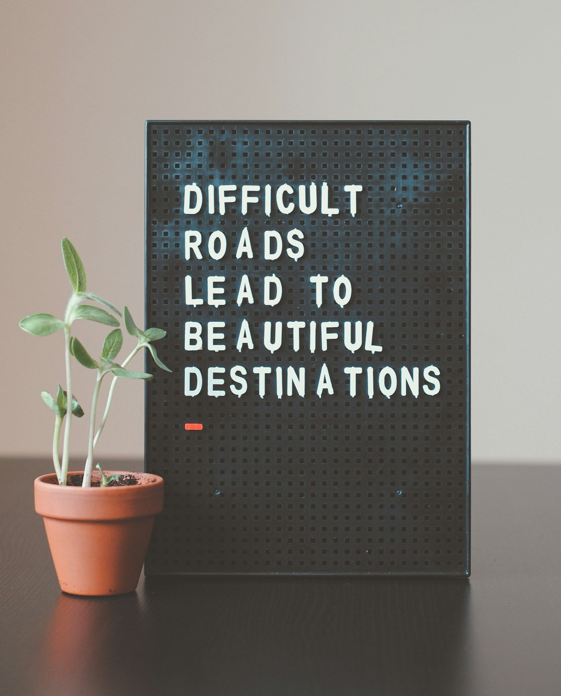 A sign with a motivational quote that reads 'Difficult roads lead to beautiful destinations' next to a potted plant on a dark surface against a plain background.