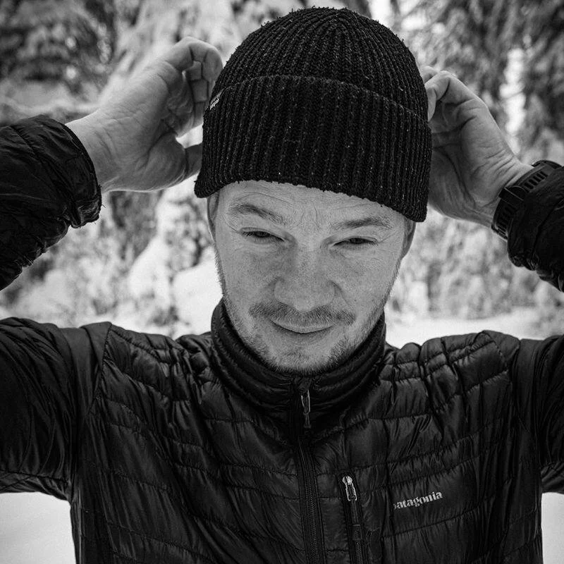 Jukka Tallinen outdoors in a winter setting, adjusting a knitted beanie on his head, wearing a puffer jacket.