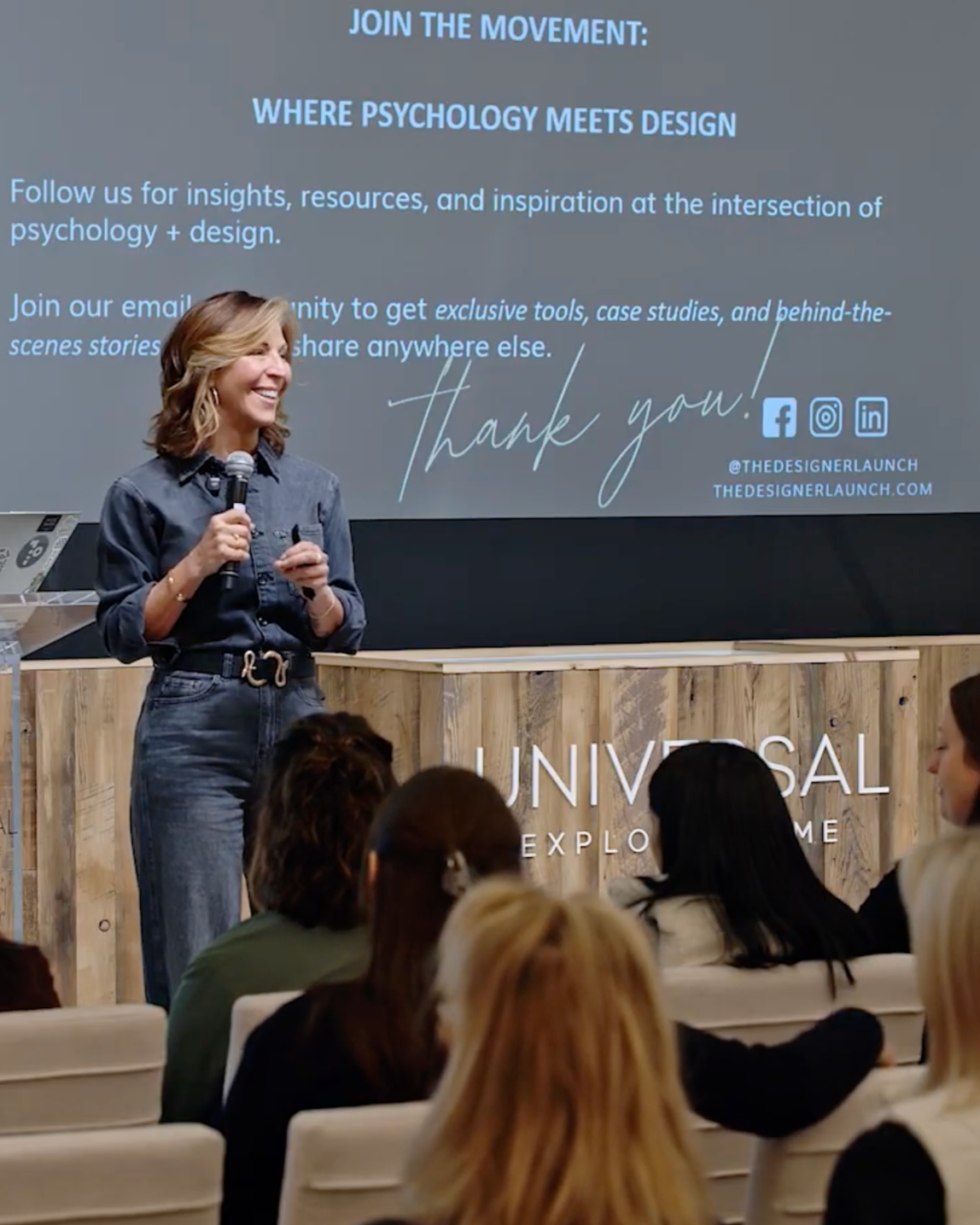 A woman holding a microphone speaking in front of a large screen at a presentation or conference. The screen displays text about joining a movement where psychology meets design, with branding for Universal and social media icons.