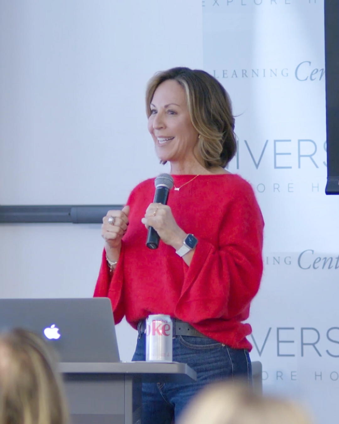 A woman in a red sweater speaking into a microphone during a presentation at a learning center. There is a laptop and a Diet Coke can on a table in front of her.