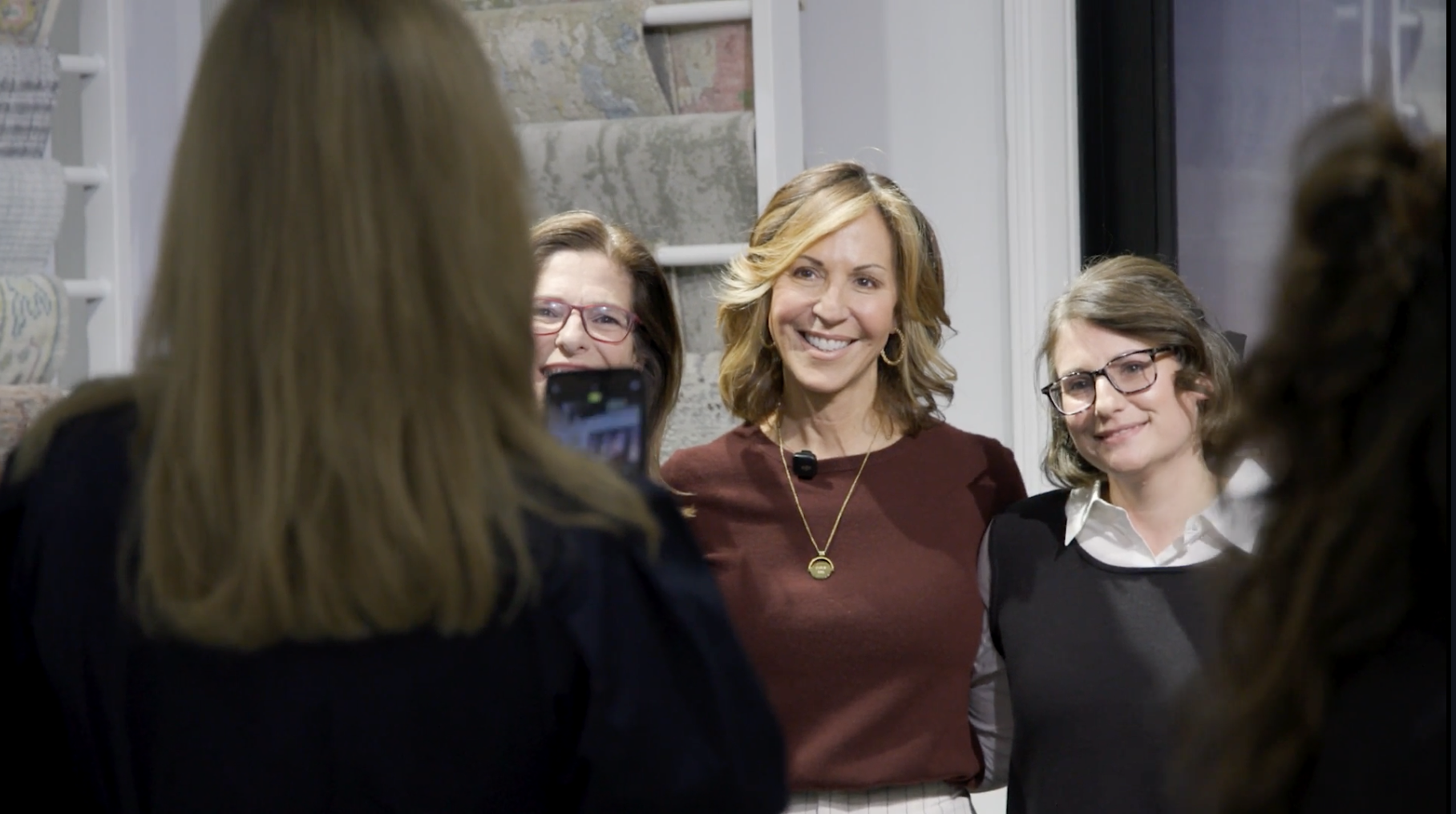 A group of women taking a photograph or selfie together inside a store, with one woman holding a phone, and shelves with carpet or rug samples in the background.