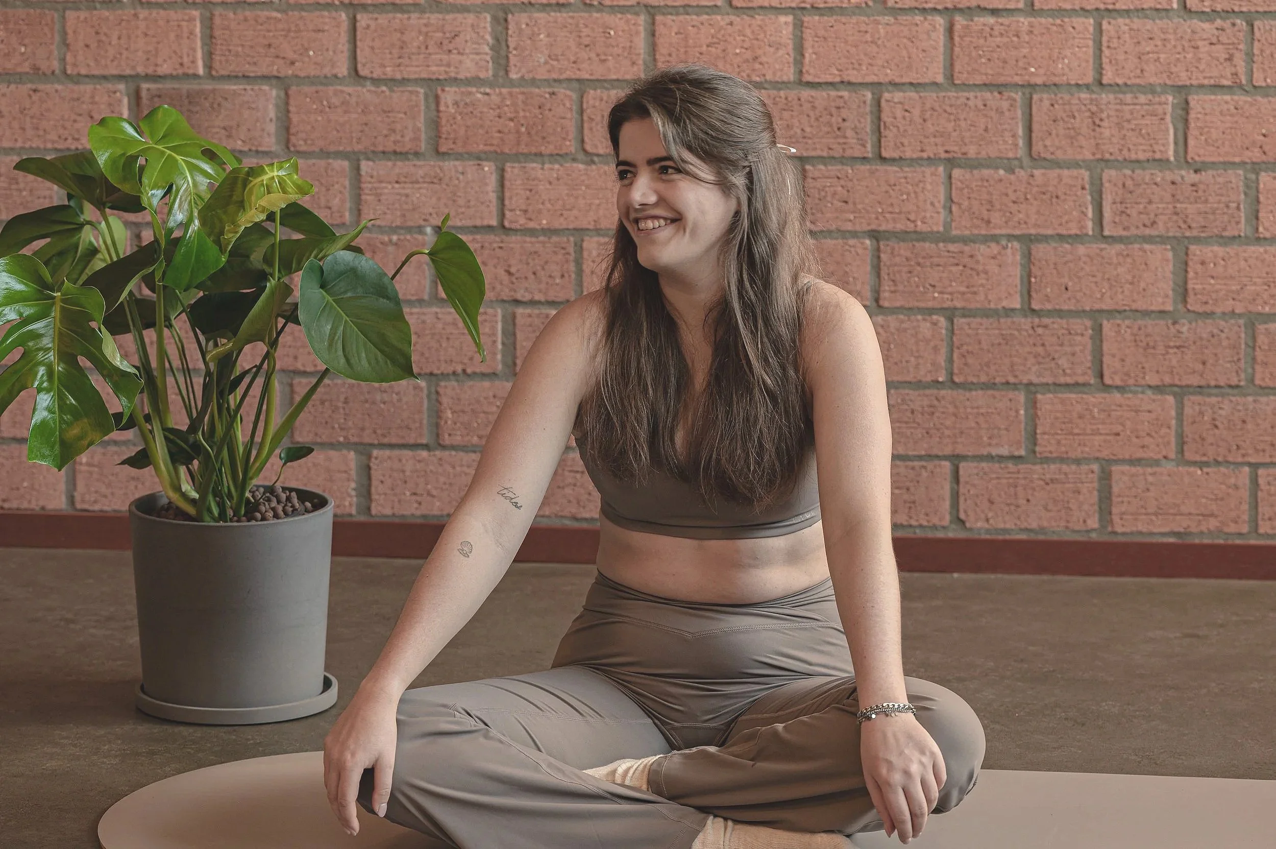 vrouw die op een mat zit in een yogakleding, glimlachend, met een plant op de achtergrond tegen een bakstenen muur.