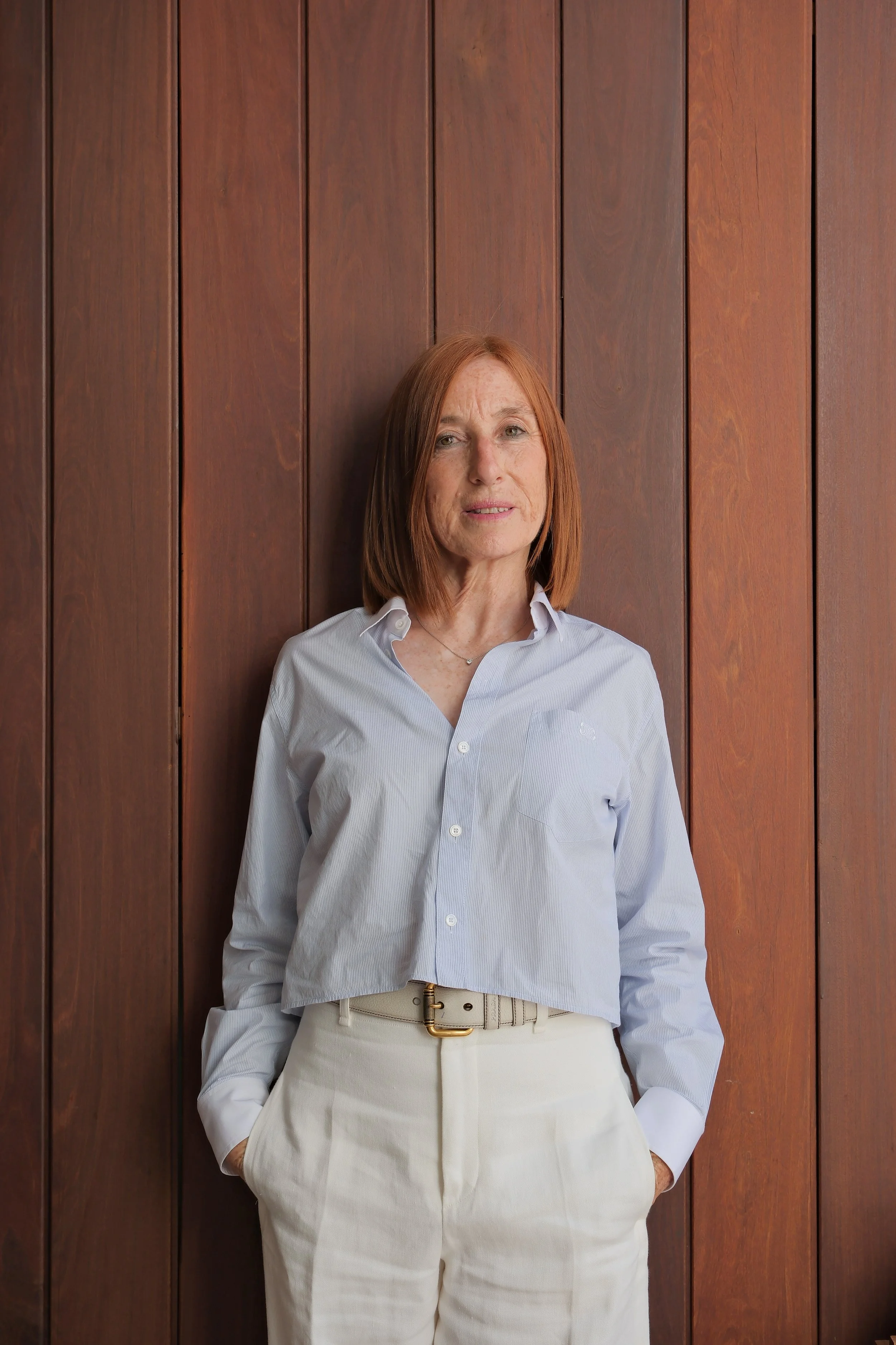 Woman in white shirt and beige pants, brown hair, standing against a wooden wall.