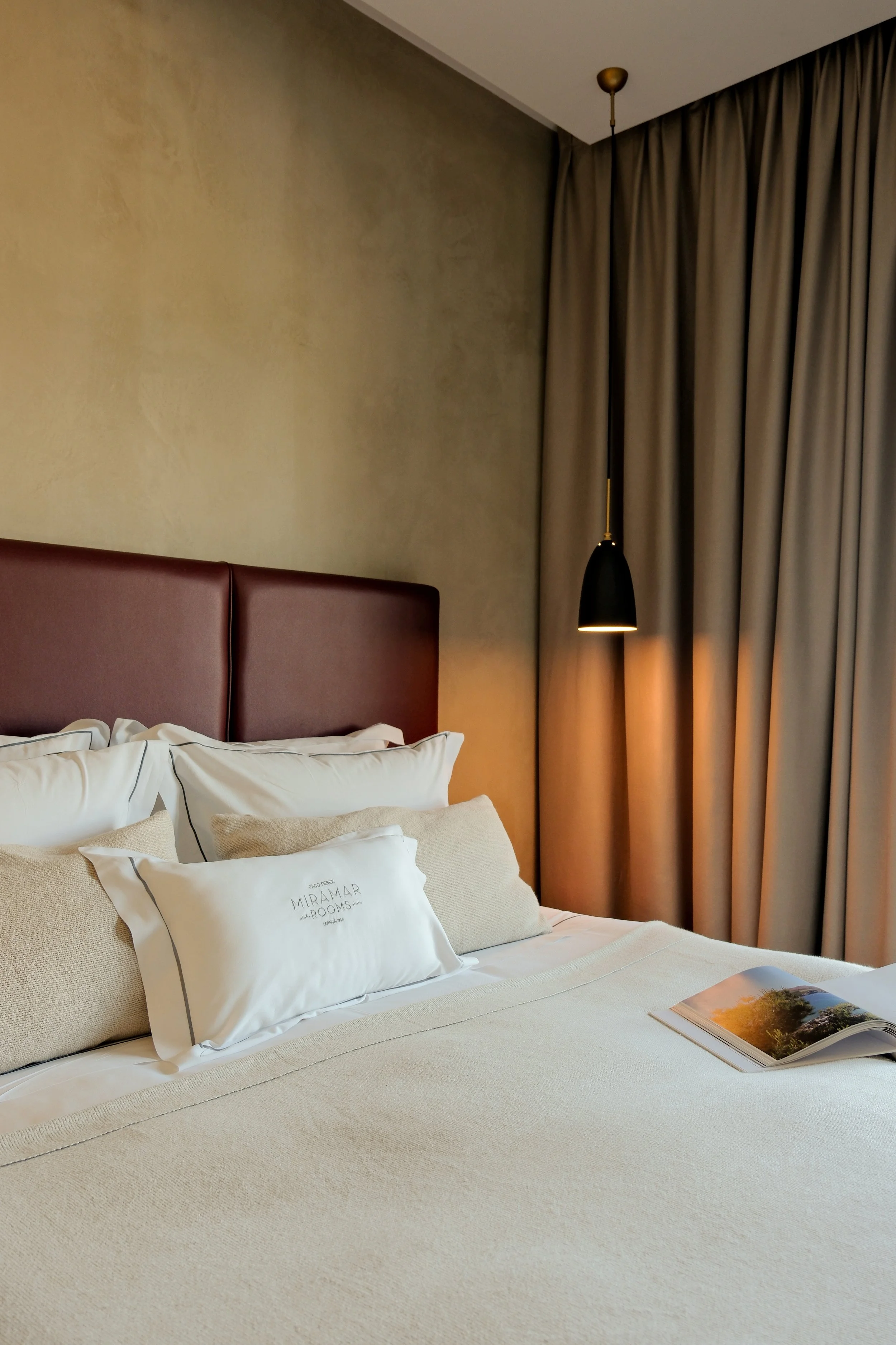 Hotel bed with white and beige pillows, table with magazine, gray curtains and black hanging lamp.