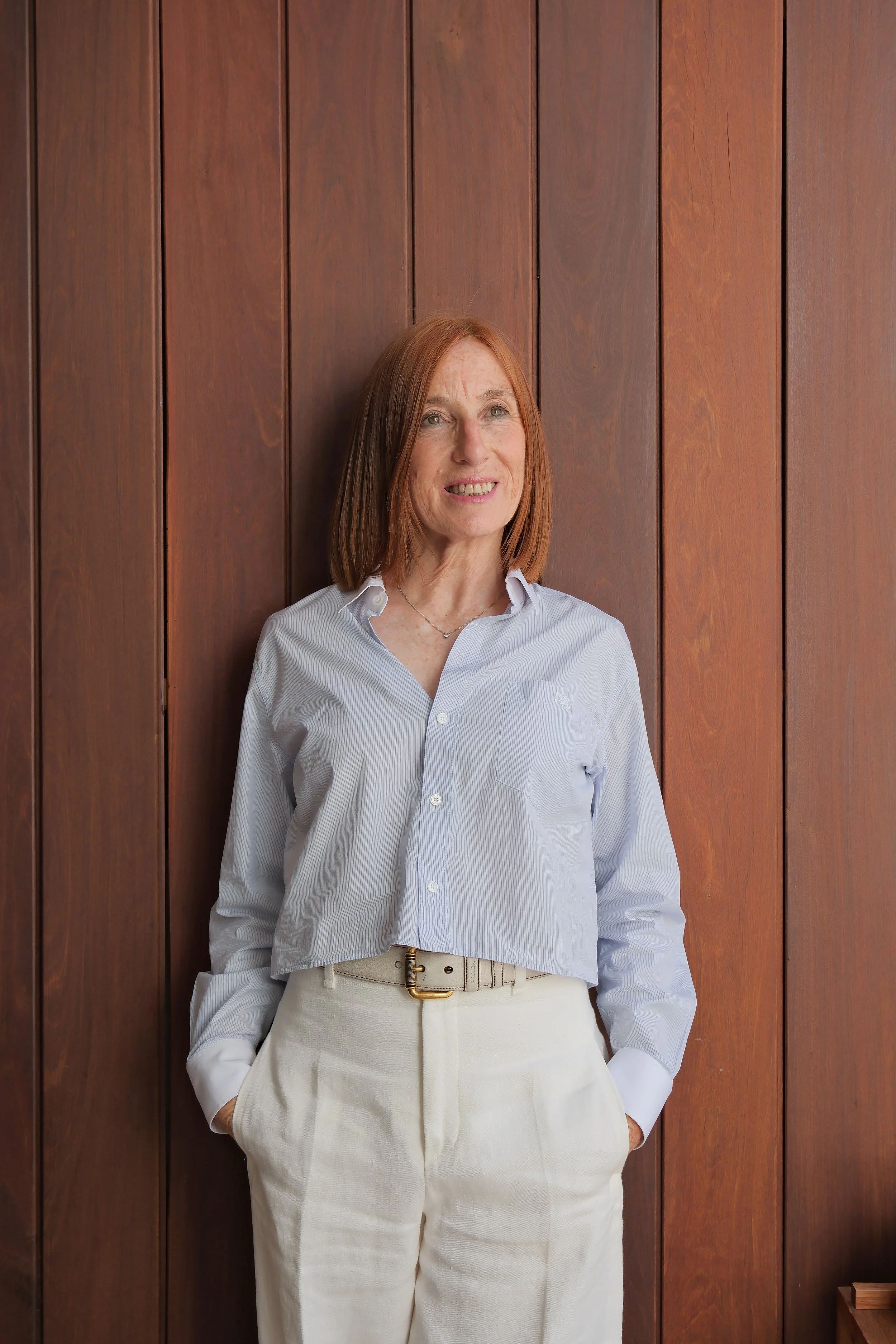 Light brown-haired woman in white blouse and beige pants, standing next to wooden wall.