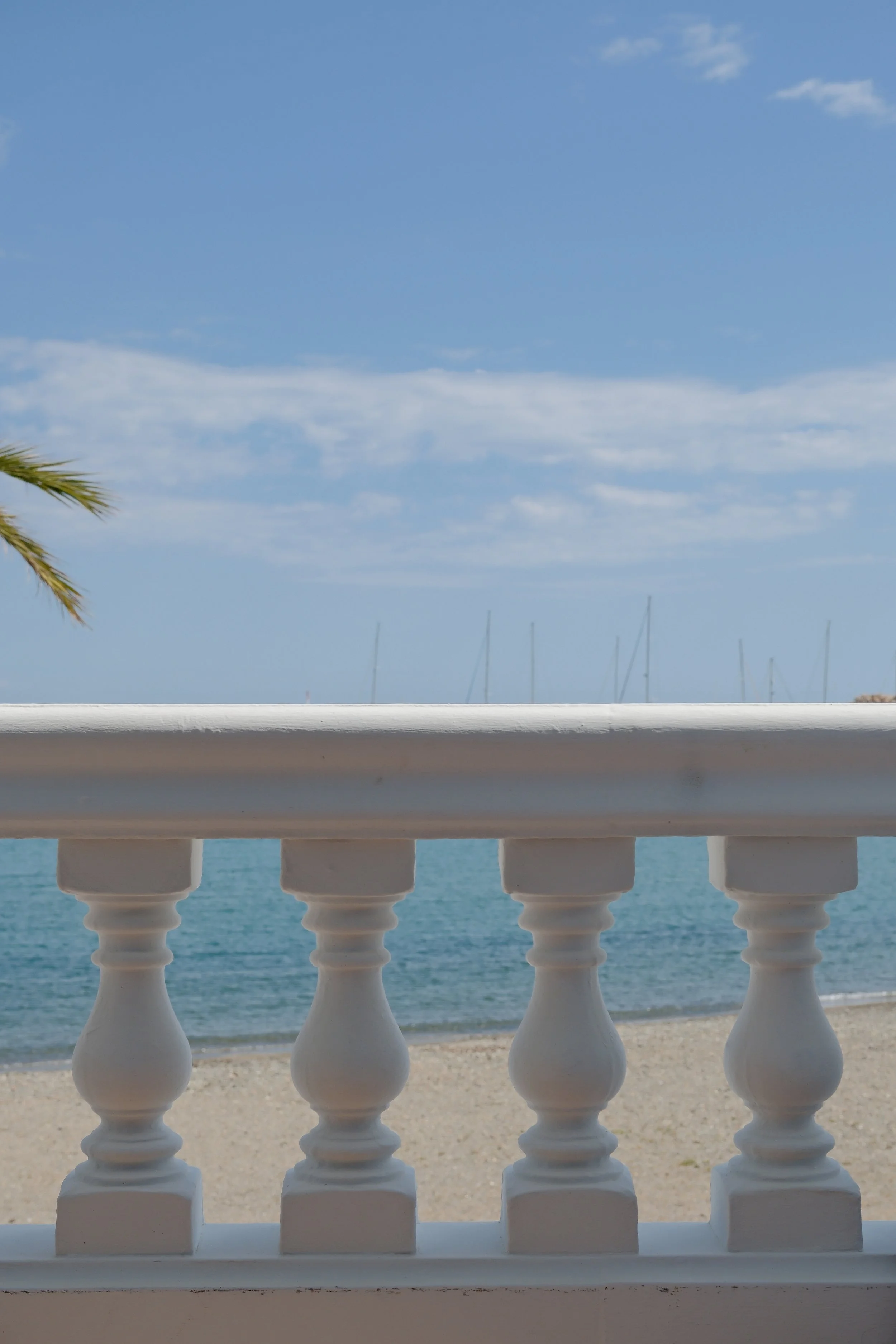 View of the sea from a terrace with white painted wooden railing, sand on the beach, and boat sails on the horizon under a partly cloudy sky.