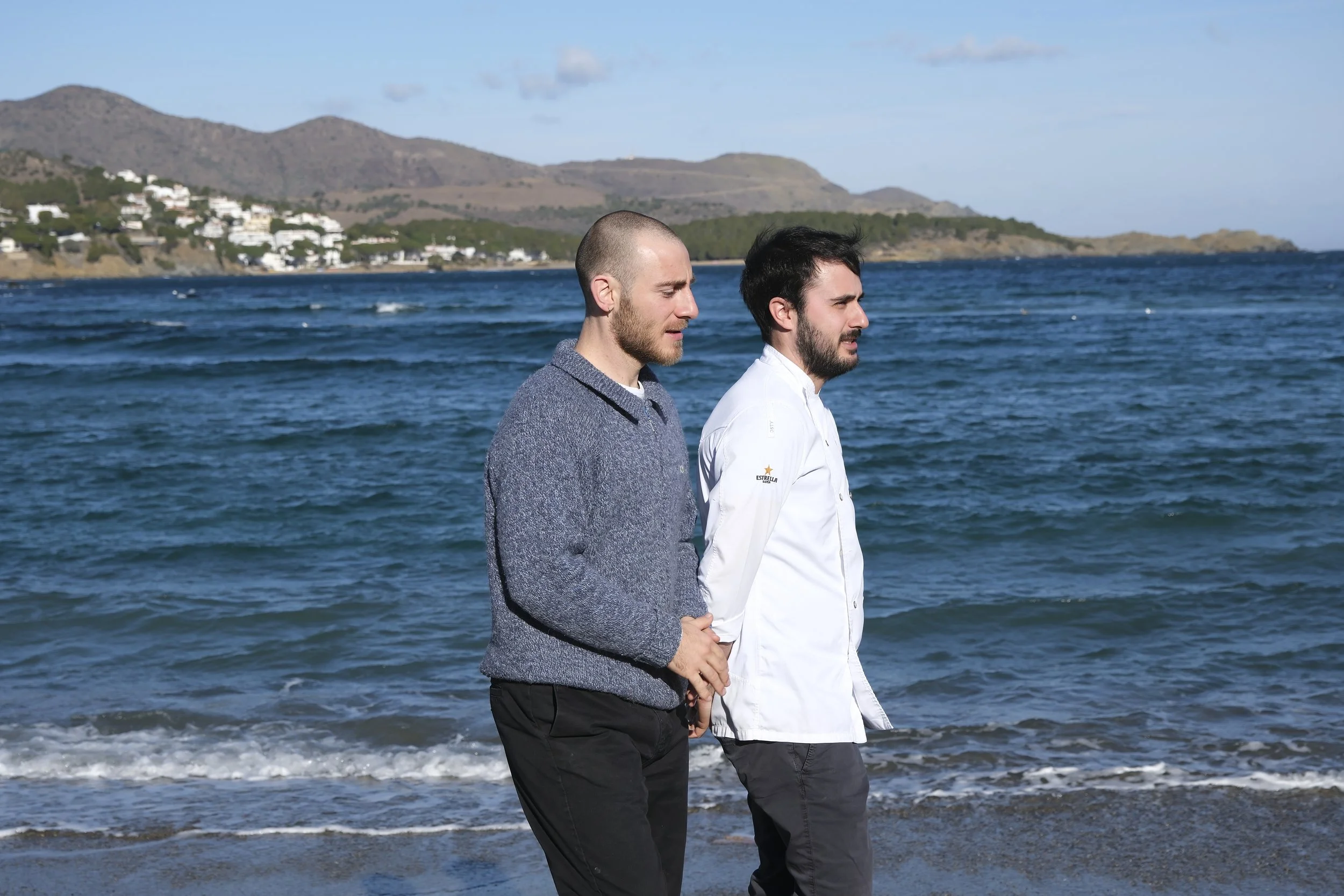 Two men walking along the seashore with mountains and white houses in the background, on a sunny day with clear skies.