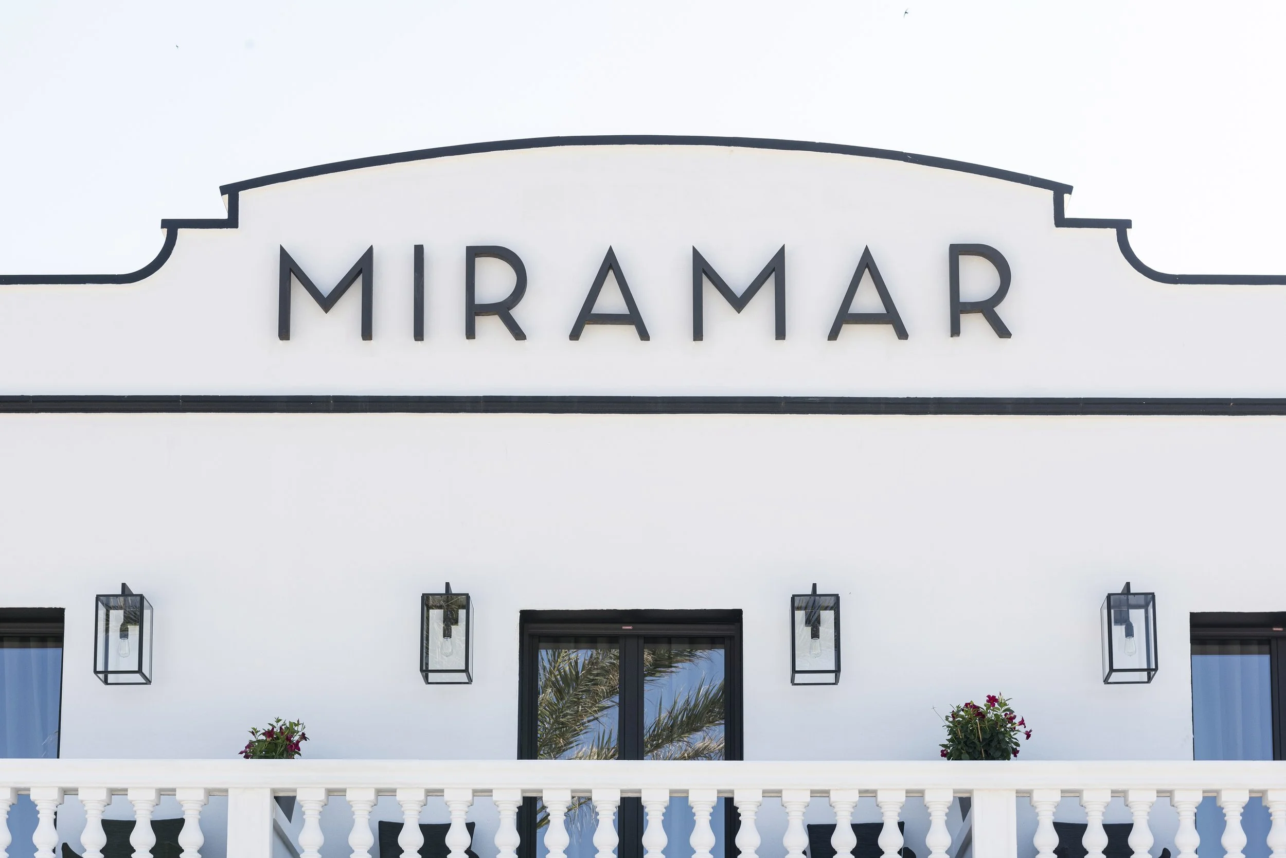 Fachada de un edificio con la palabra 'MIRAMAR' en letras grandes y negras, con ventanas y sillas blancas en un balcón, decorado con flores.