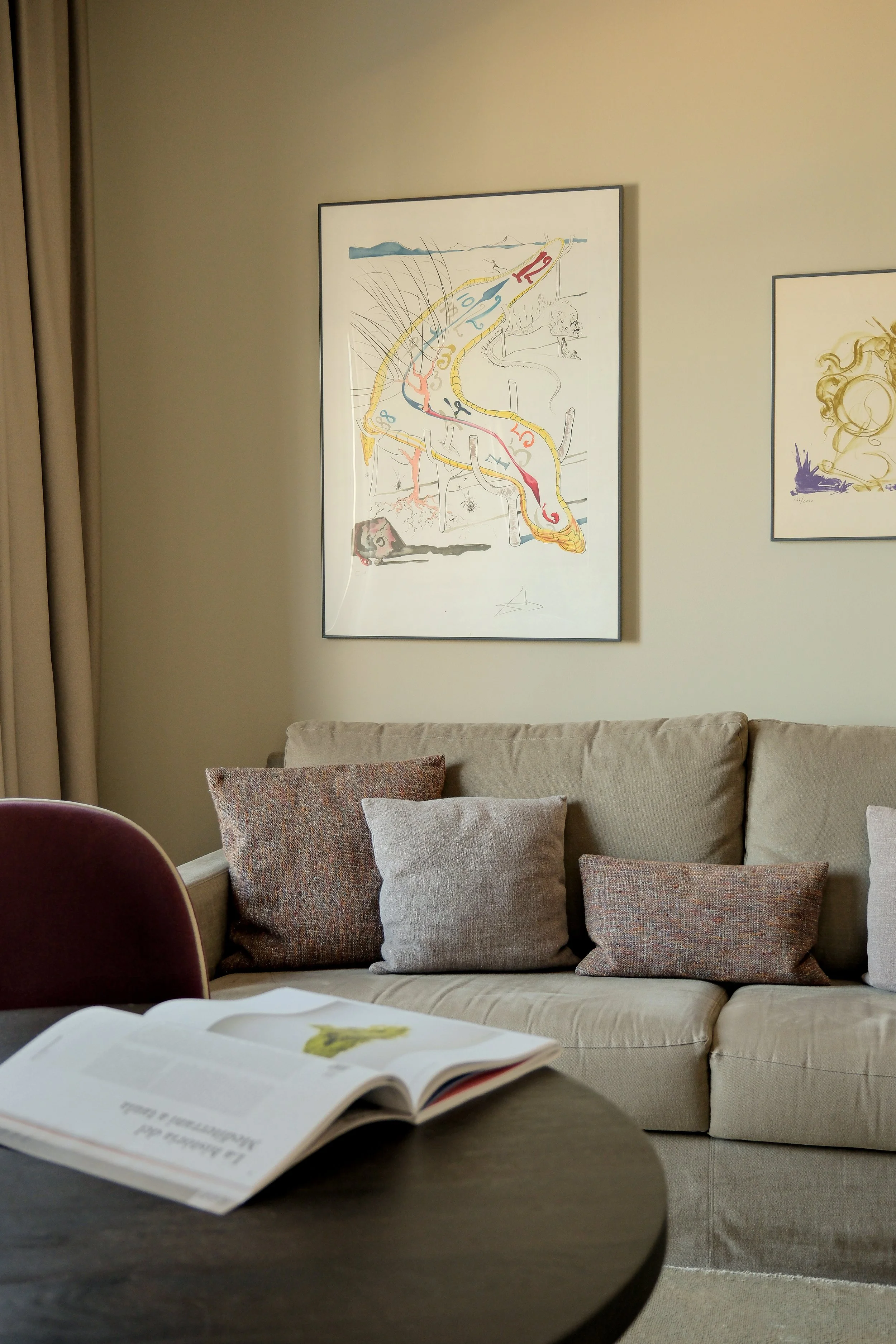 Living room with a beige armchair, cushions and pictures on the wall, near a table with an open book.