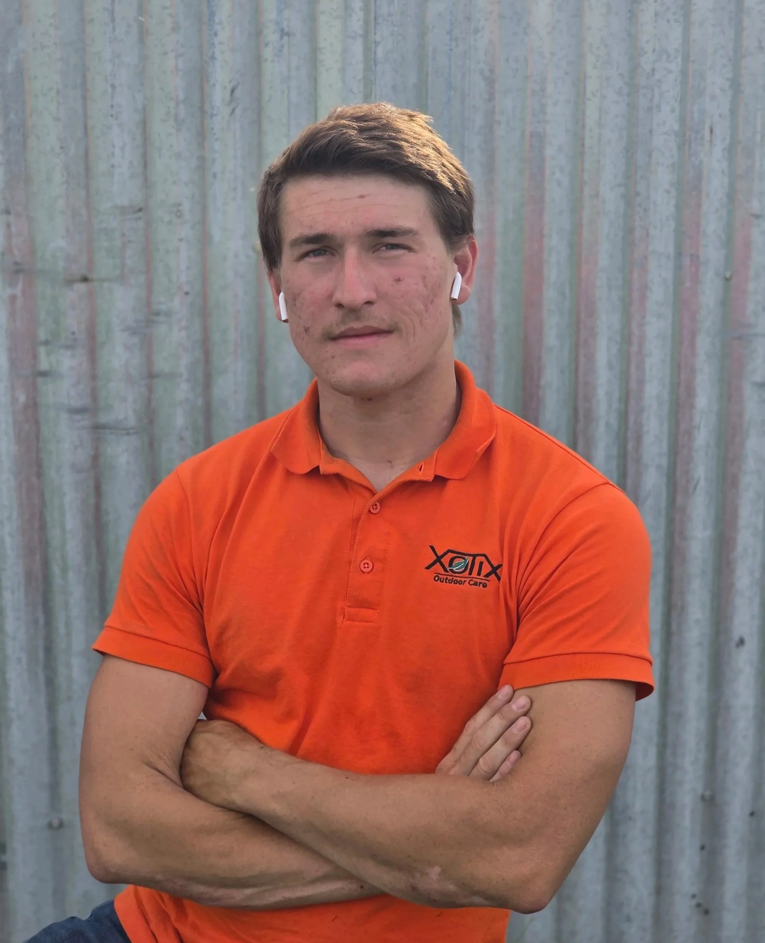 A young man with short brown hair and blue eyes standing outdoors in front of a corrugated metal wall. He is wearing white wireless earbuds, an orange polo shirt with a logo that reads 'Outdoor Care,' and has his arms crossed.