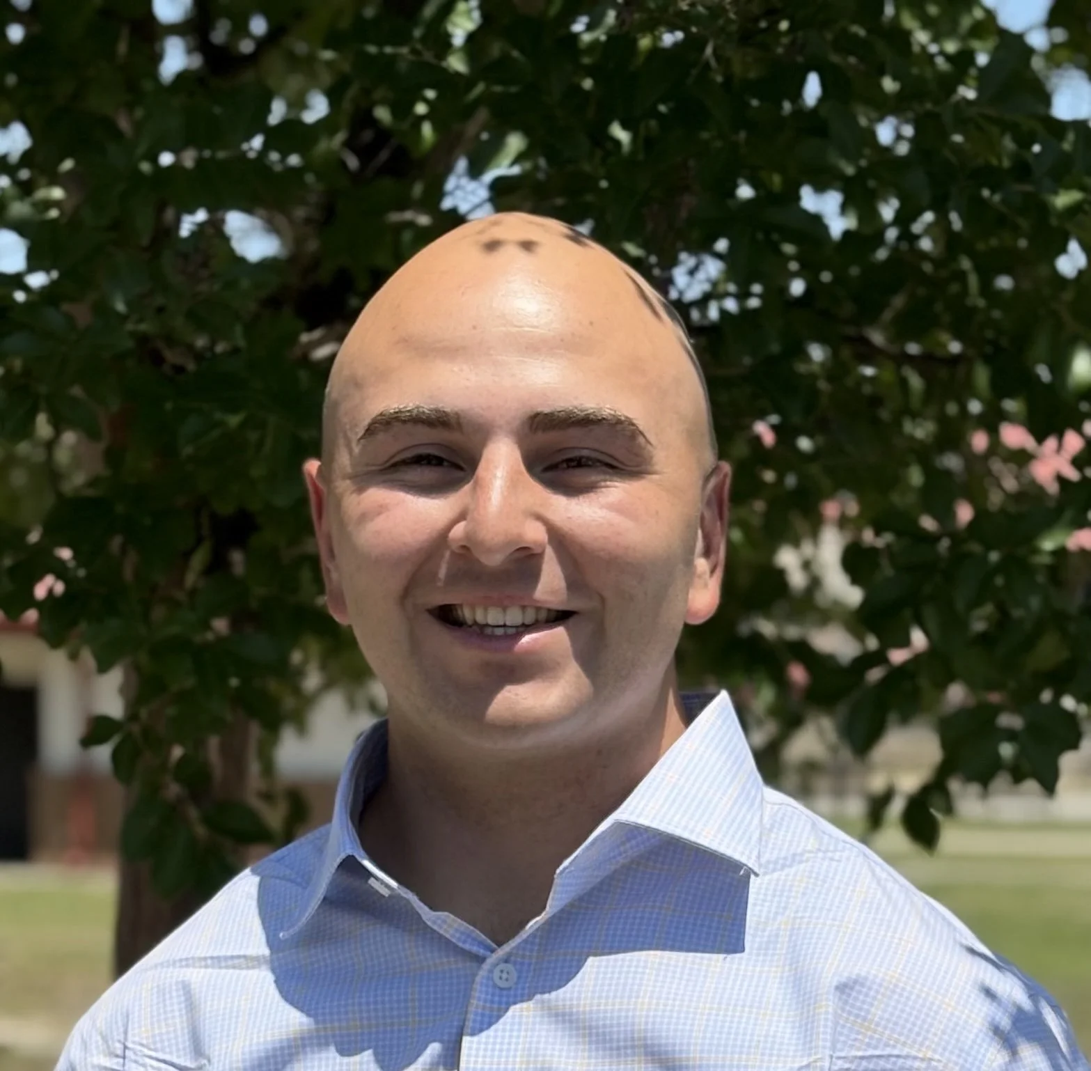 Photo of a smiling man with a shaved head wearing a blue striped shirt, standing outdoors in front of a tree with pink flowers and green leaves.