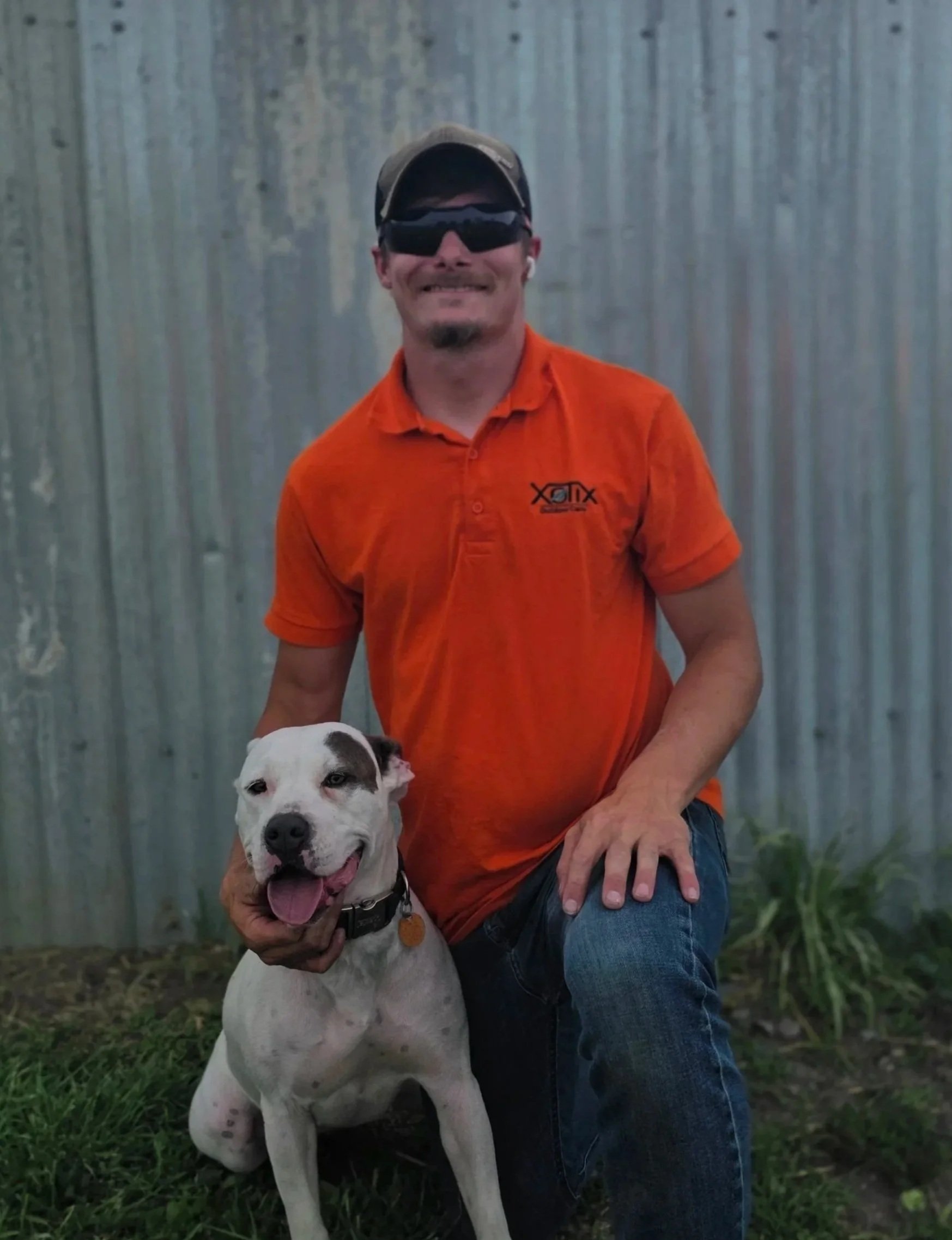 A man in an orange shirt and blue jeans kneeling outdoors with his arm around a large white and black dog. The man is wearing a cap and dark sunglasses, and the background is a weathered metal fence surrounded by greenery.