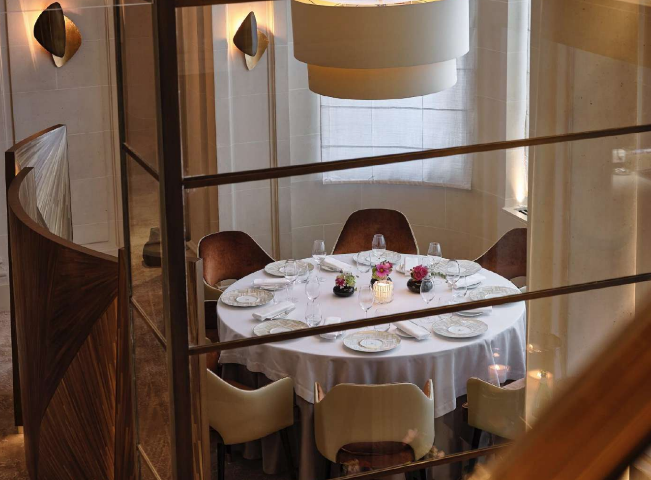 Salle à manger élégante avec une grande table ronde ornée d'une nappe blanche, de fleurs et de verres, entourée de chaises en velours marron et crème, vue à travers une grille de verre.