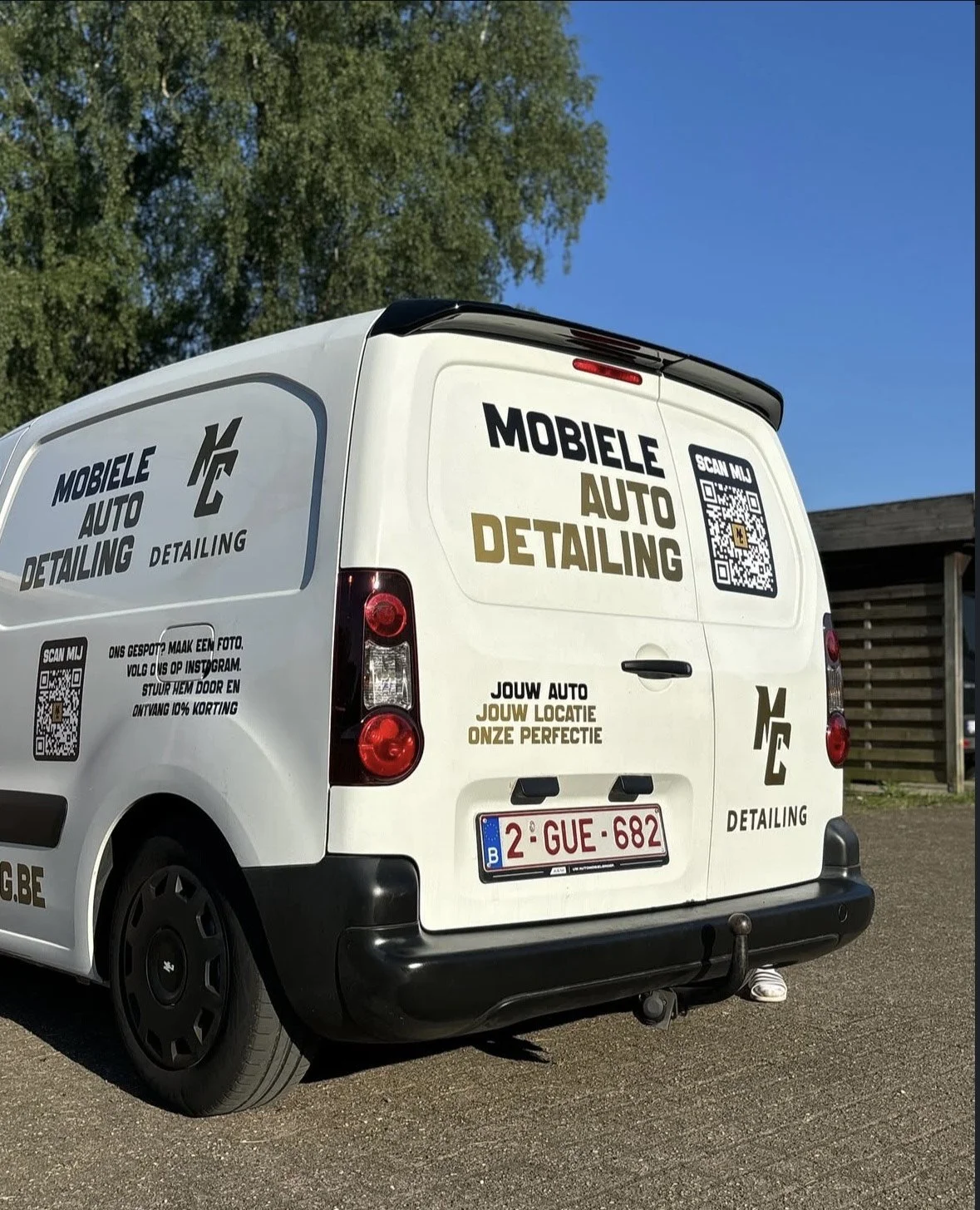 Een witte bedrijfswagen met advertenties voor auto detailing, QR-codes, een Belgisch nummerbord en een houten omheining op de achtergrond.