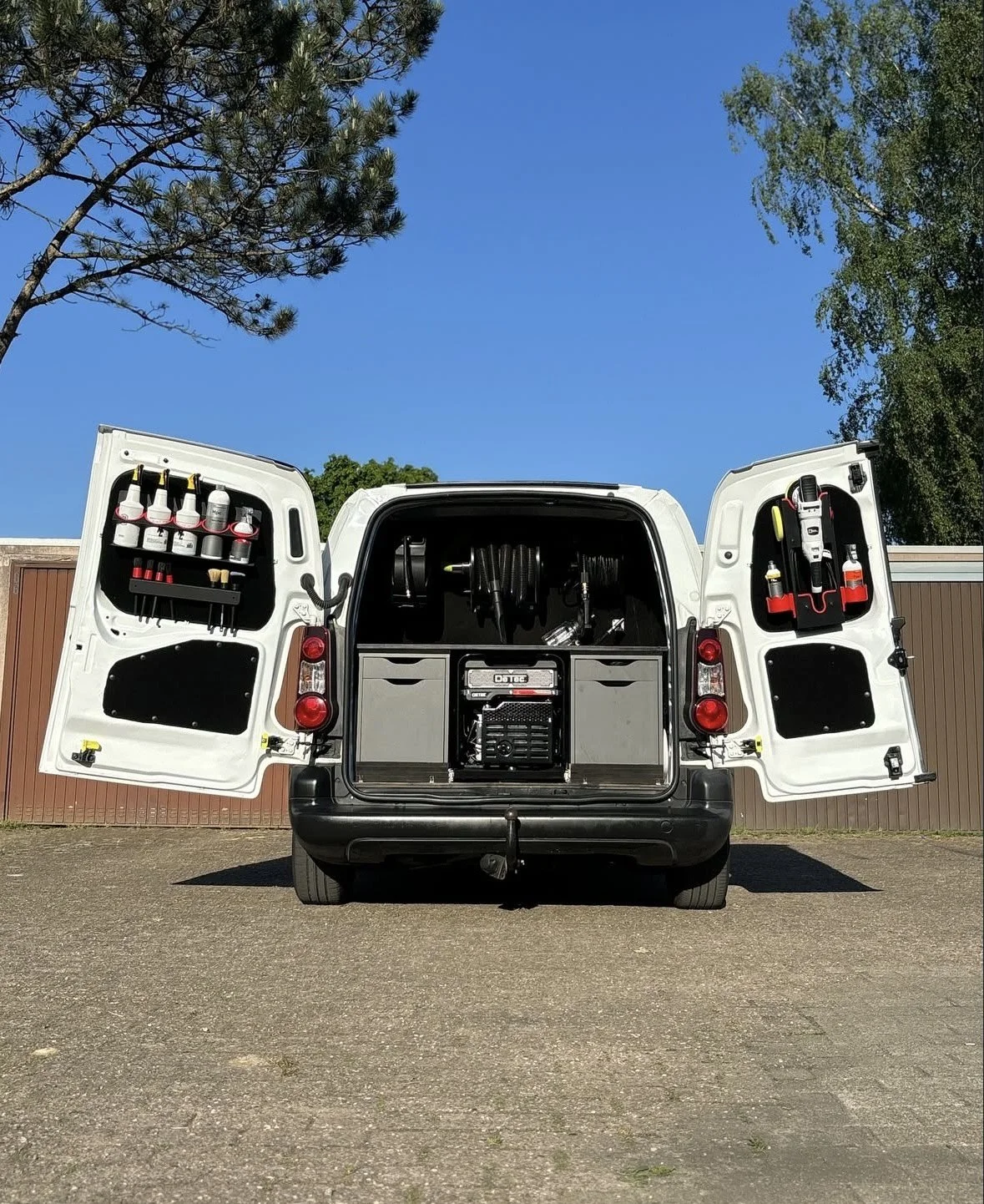Achterkant van een professioneel ingerichte werkbus met geopende deuren, gevuld met schoonmaakmiddelen, hogedrukslangen, en een generator voor mobiele autoreiniging of detailing service.
