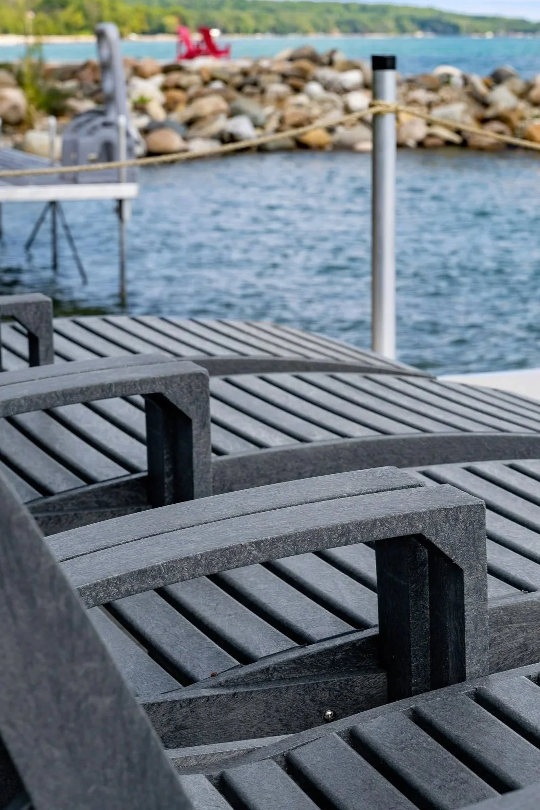 Empty gray lounge chairs on a dock by a body of water, with boats and a rocky shoreline in the background.