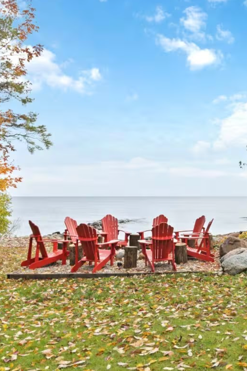 A group of red Adirondack chairs arranged in a circle around a fire pit on a rocky beach, with fallen autumn leaves on the ground and a body of water and blue sky with scattered clouds in the background.
