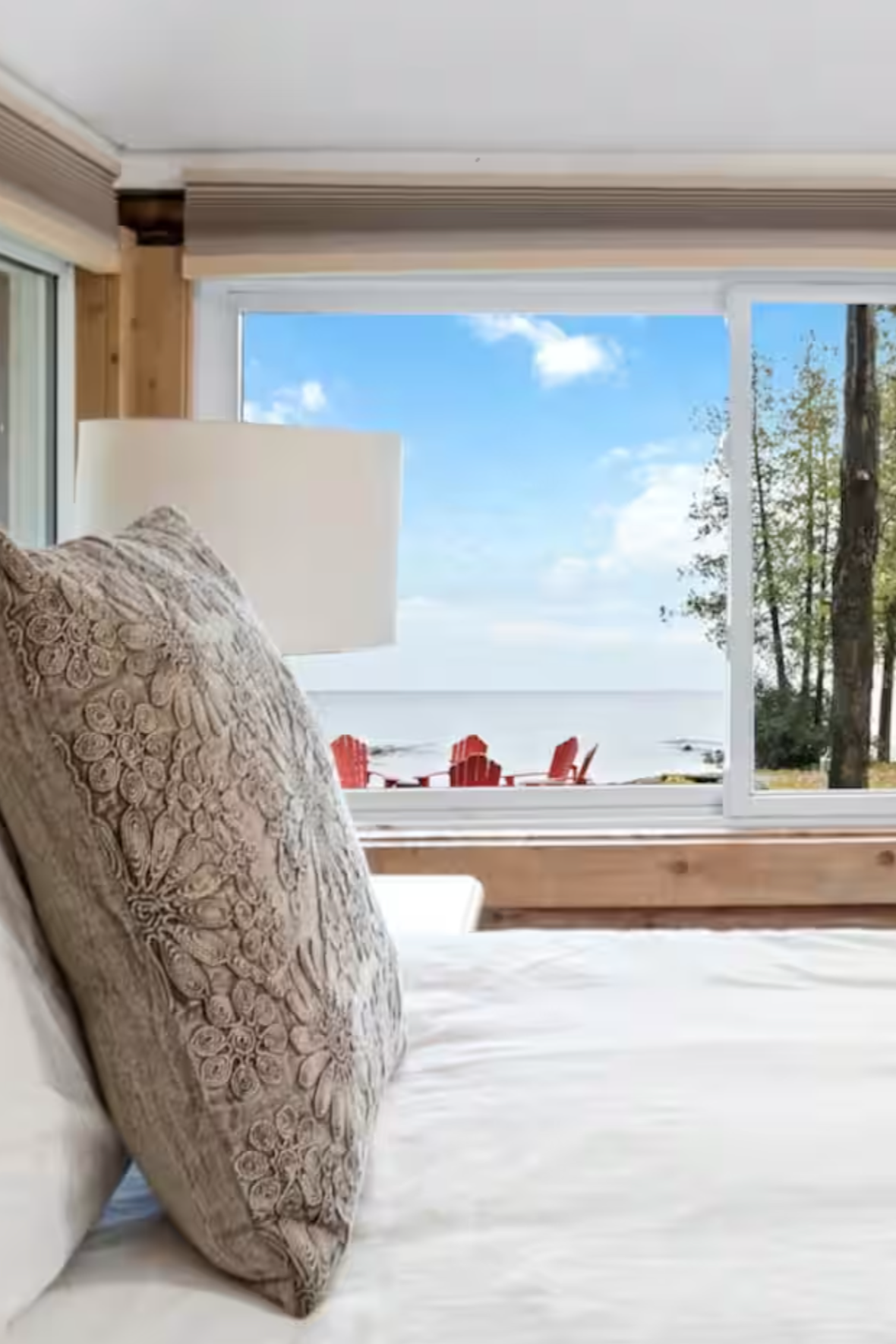 View of a bedroom with an open window showing a beach scene with red chairs, trees, and a blue sky with clouds.