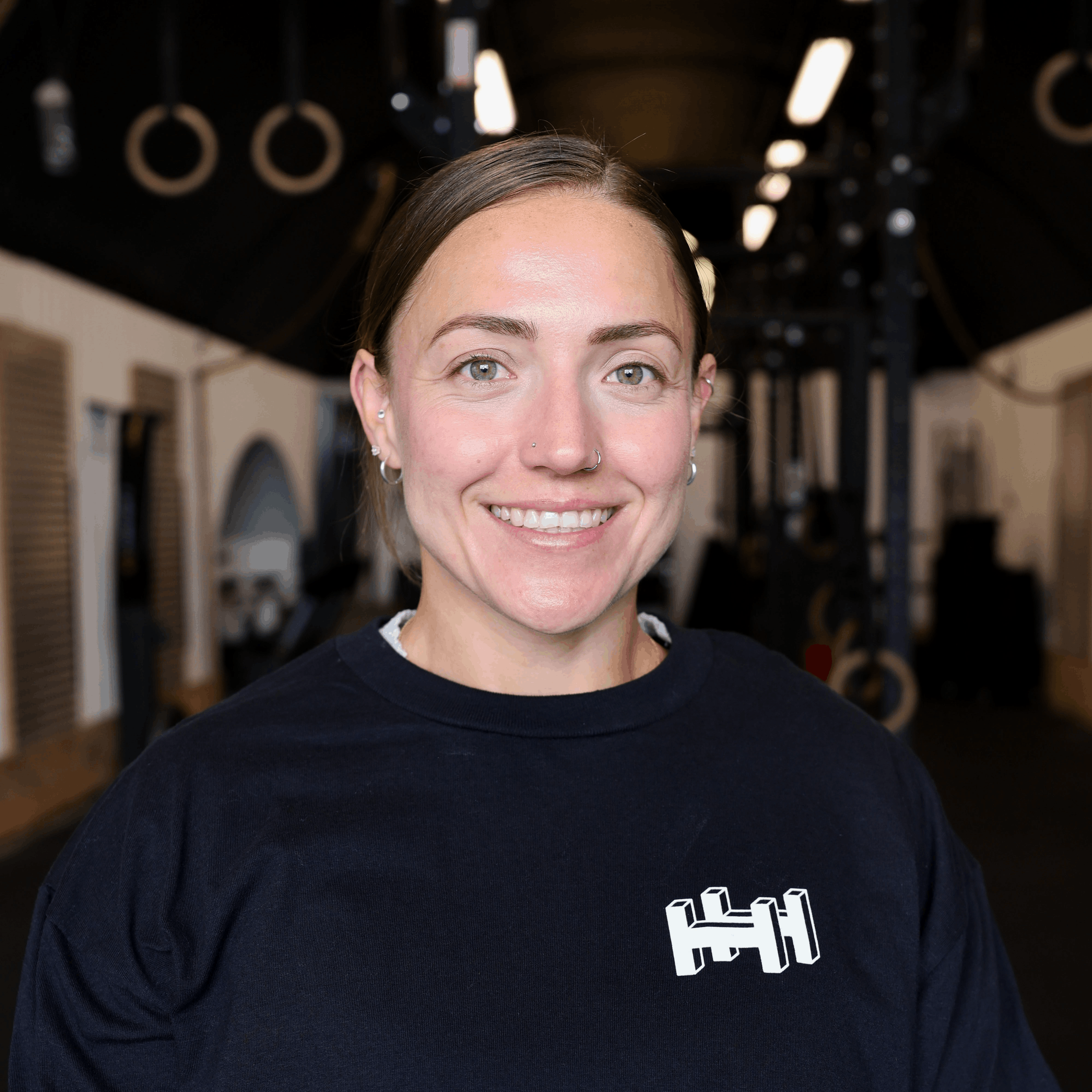 A woman with light skin, short brown hair, and blue eyes smiling at the camera in a gym or fitness studio. She is wearing a black shirt with a small white logo and a purple high-neck shirt underneath.