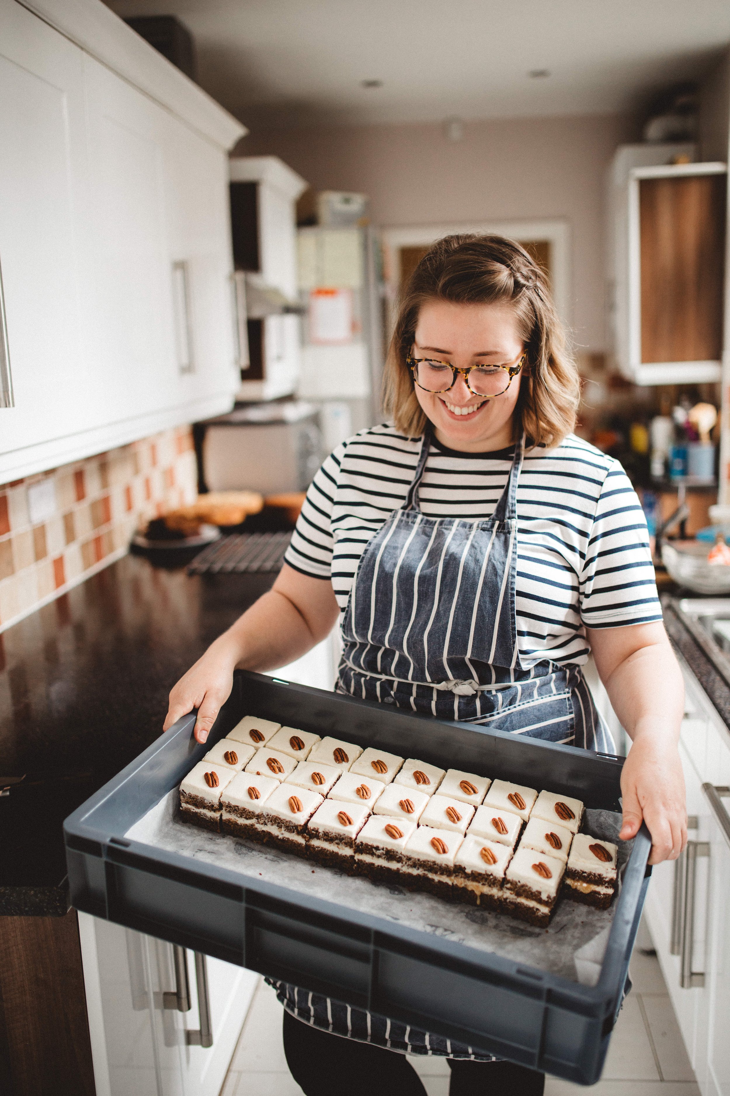 A woman with glasses and a striped shirt smiles while holding a baking tray of frosted brownies decorated with pecans in a kitchen.