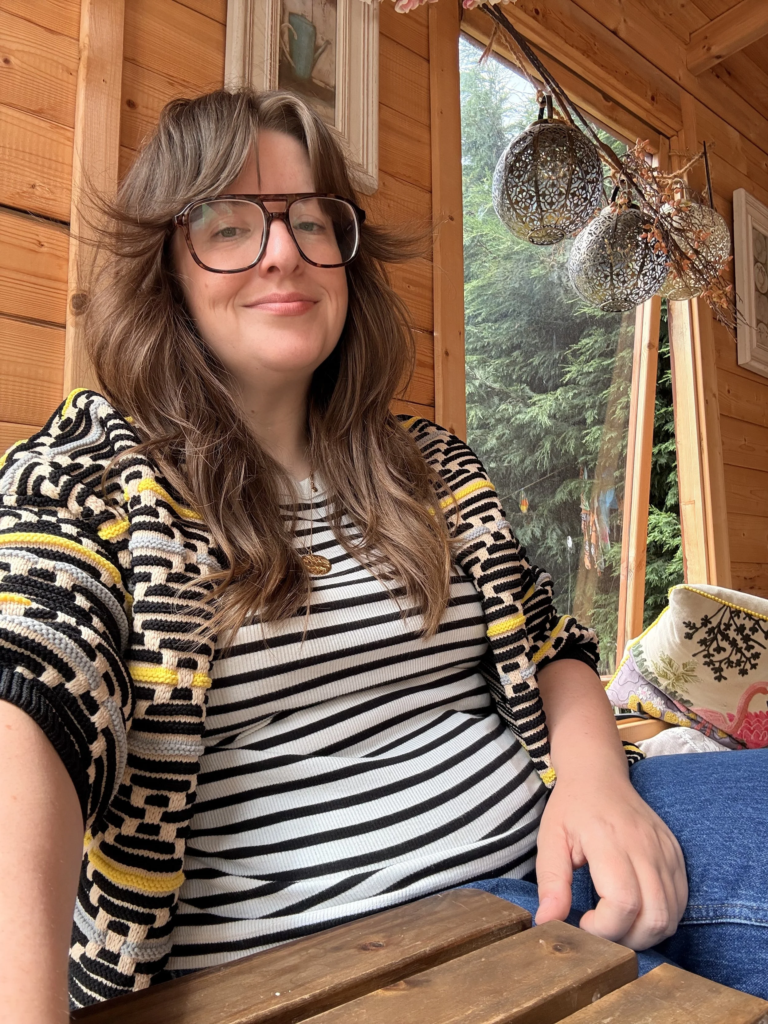 A woman with long, wavy brown hair, wearing large glasses, a striped shirt, and a multicolored crochet cardigan, points with her finger at a wooden table inside a cozy, wood-paneled room with a large window showing outdoors greenery.