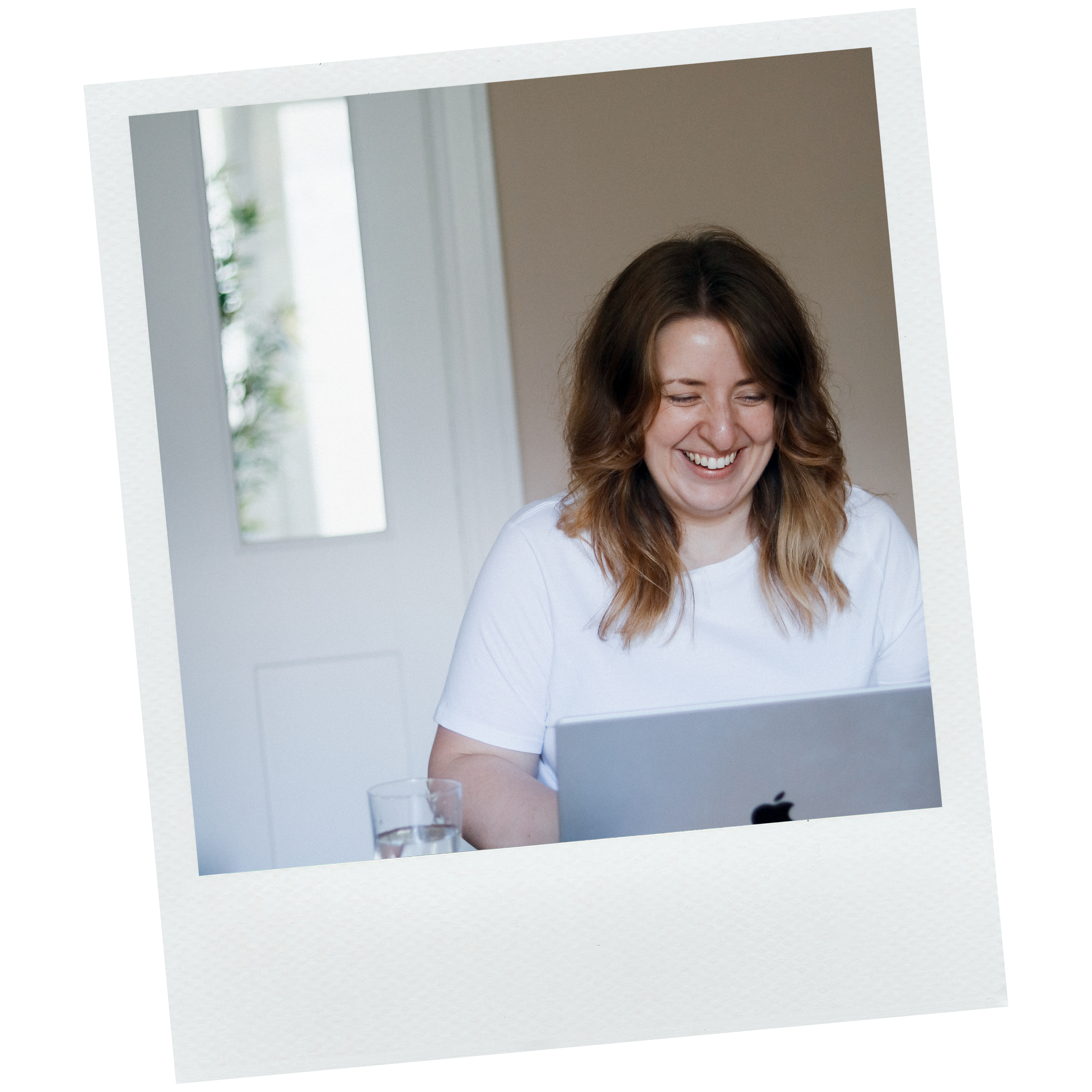 Smiling woman with brown hair and white shirt looking down in a bright room with a window and a glass of water on the table.