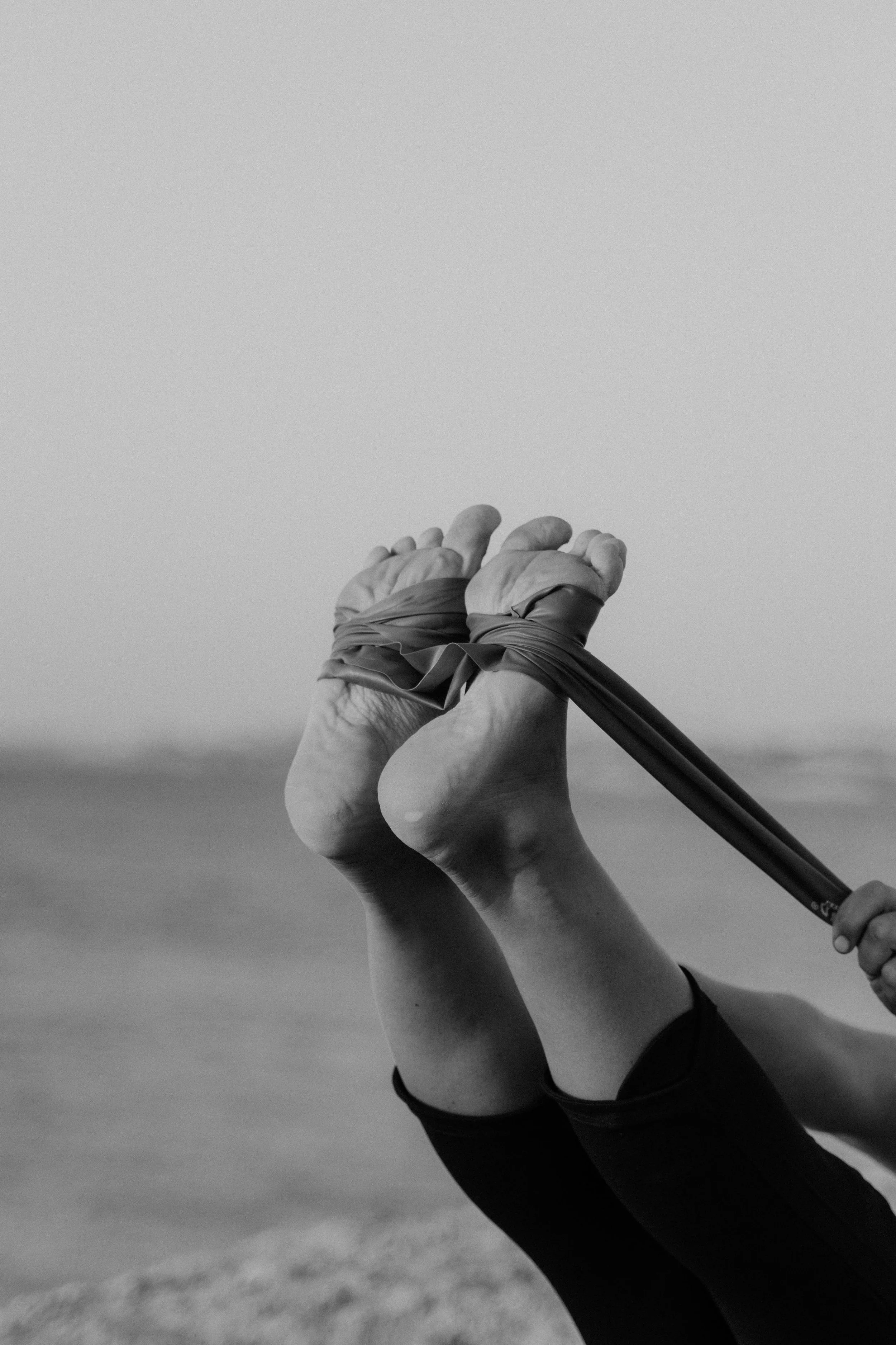 Black and white photo of a person's feet bound together with a cloth, holding a stick, with a blurred background of a beach or open land.