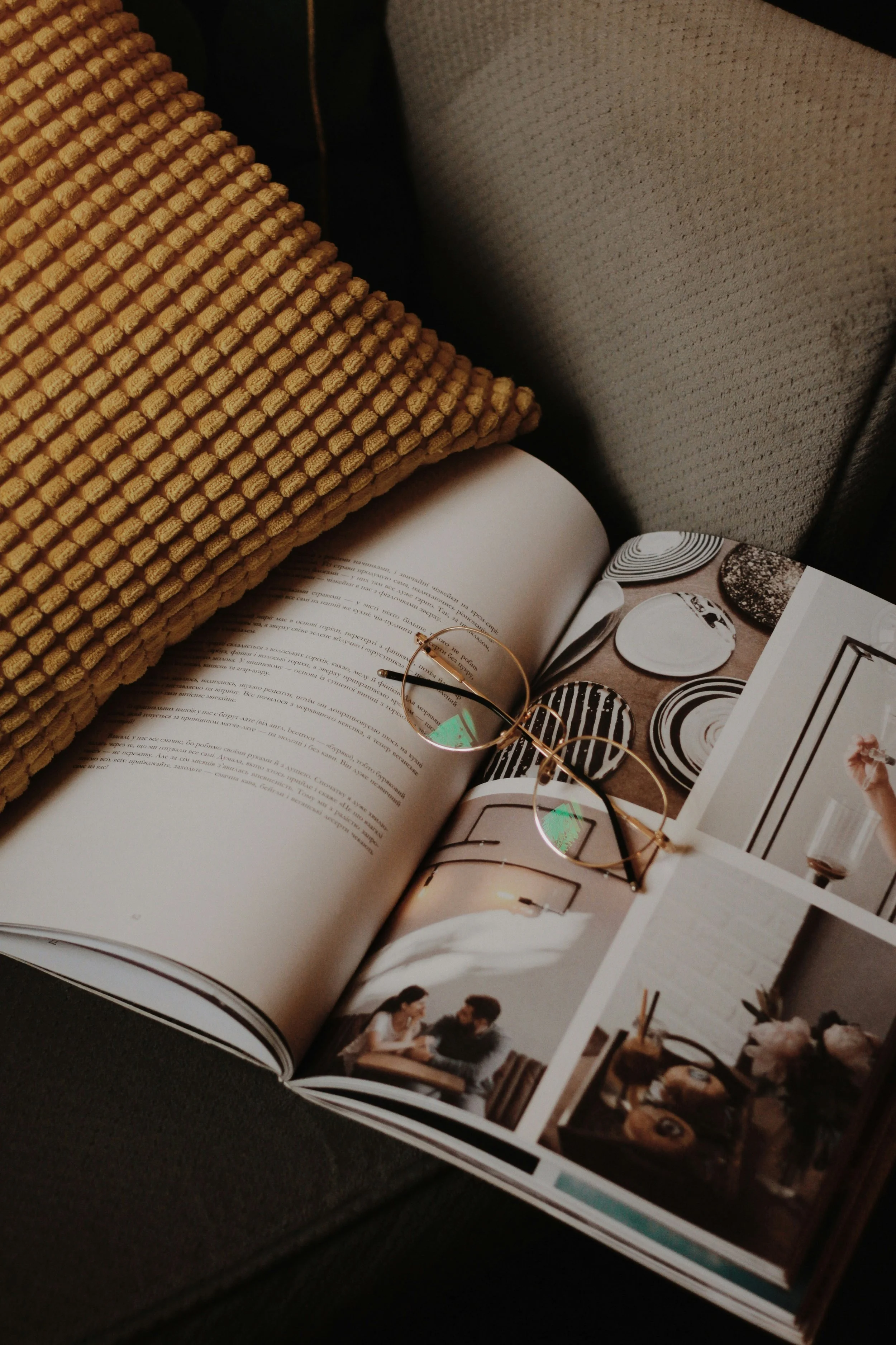 Open magazine on dark surface, showing interior design photos, a pair of gold-rimmed glasses, and a mustard-colored textured pillow.