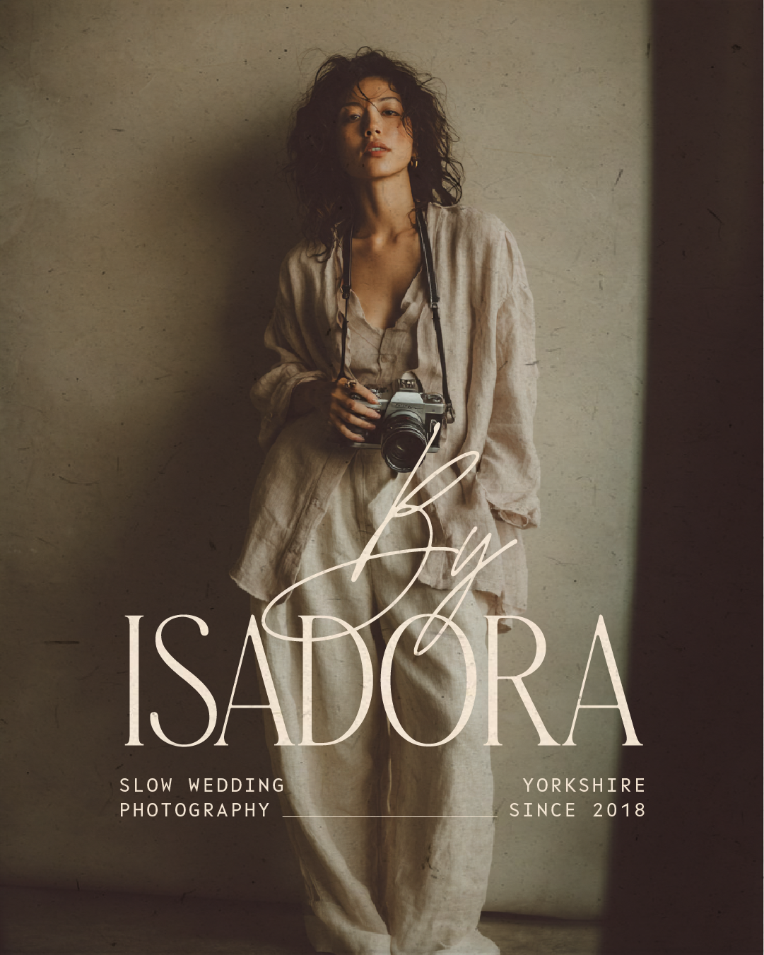 A woman with curly hair standing against a textured wall, holding a camera around her neck, wearing beige clothing, with the text 'By ISADORA' and 'Slow Wedding Photography' on the image.