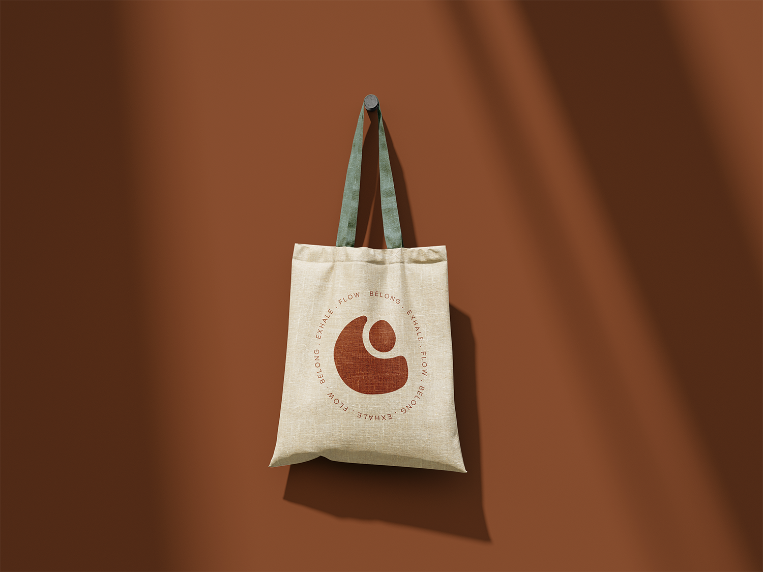 Beige tote bag hanging on a wall with green straps and a circular logo that says "Exhale. Flow. Belong." around a stylized coffee cup icon, casting a shadow on a brown wall.