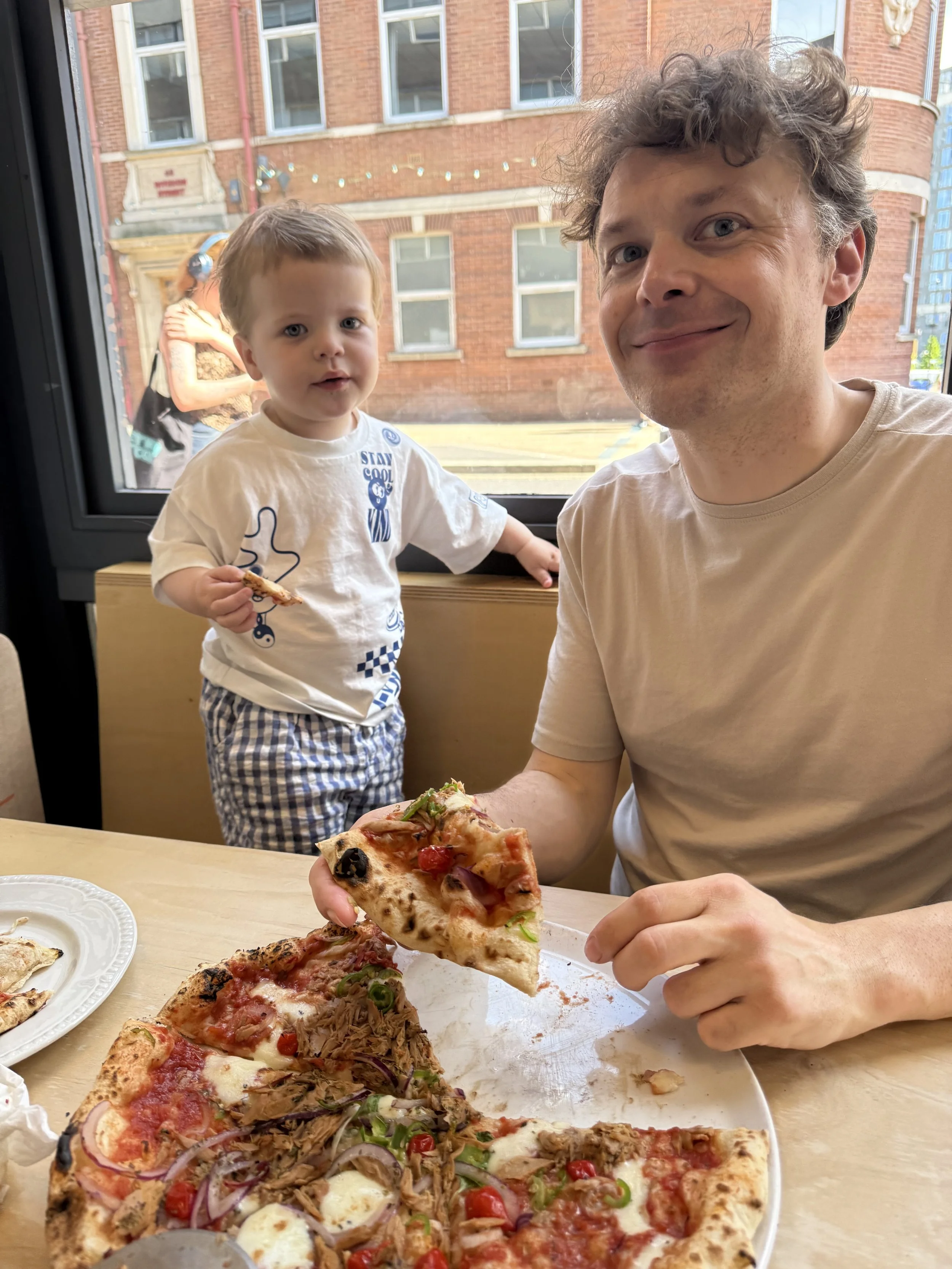 A man and a young boy sitting at a table with pizza inside a restaurant. The man is holding a slice of pizza, and the boy is standing near a window, holding a small piece of pizza and looking at the camera. The pizza on the table has various toppings, including cherry tomatoes, onions, and olives. The background outside the window shows a brick building and a woman walking by.
