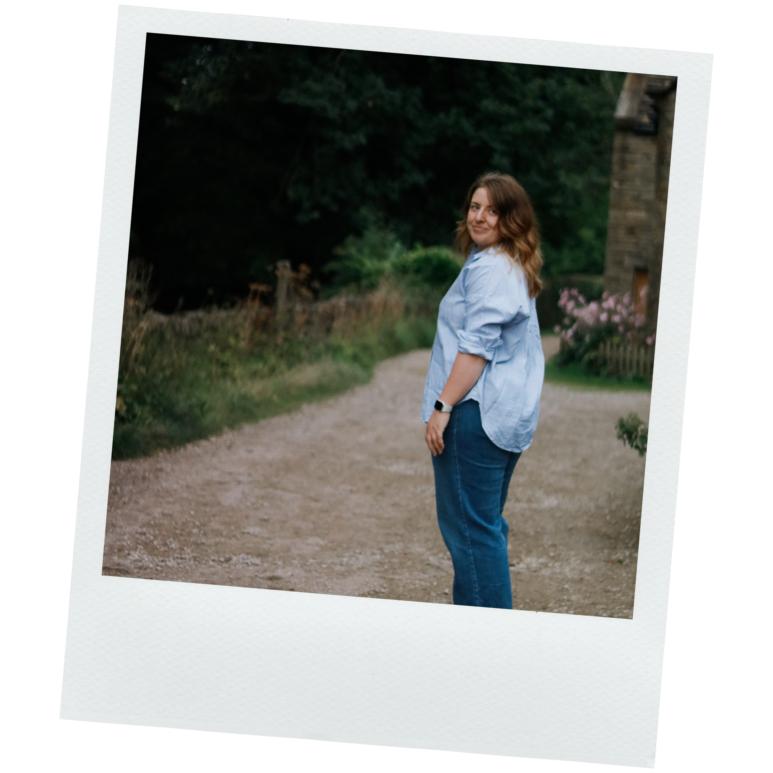 Young woman with shoulder-length brown hair wearing a light blue shirt and jeans, smiling and standing outdoors in a park or garden setting with greenery and a stone structure in the background.