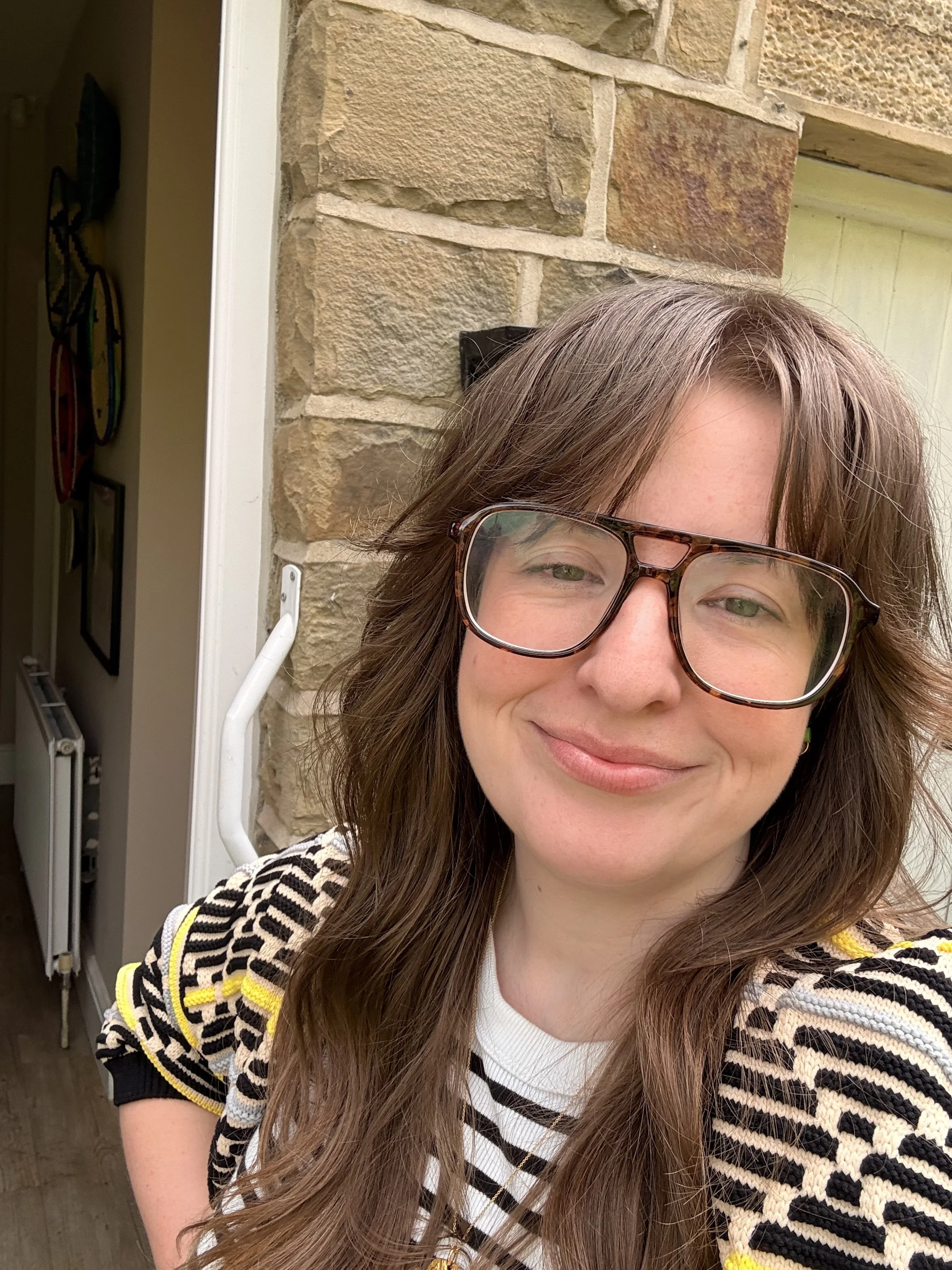 Partial view of a woman with brown hair, wearing large eyeglasses and a striped black, white, and yellow sweater, standing outside near a stone wall and a house entrance.